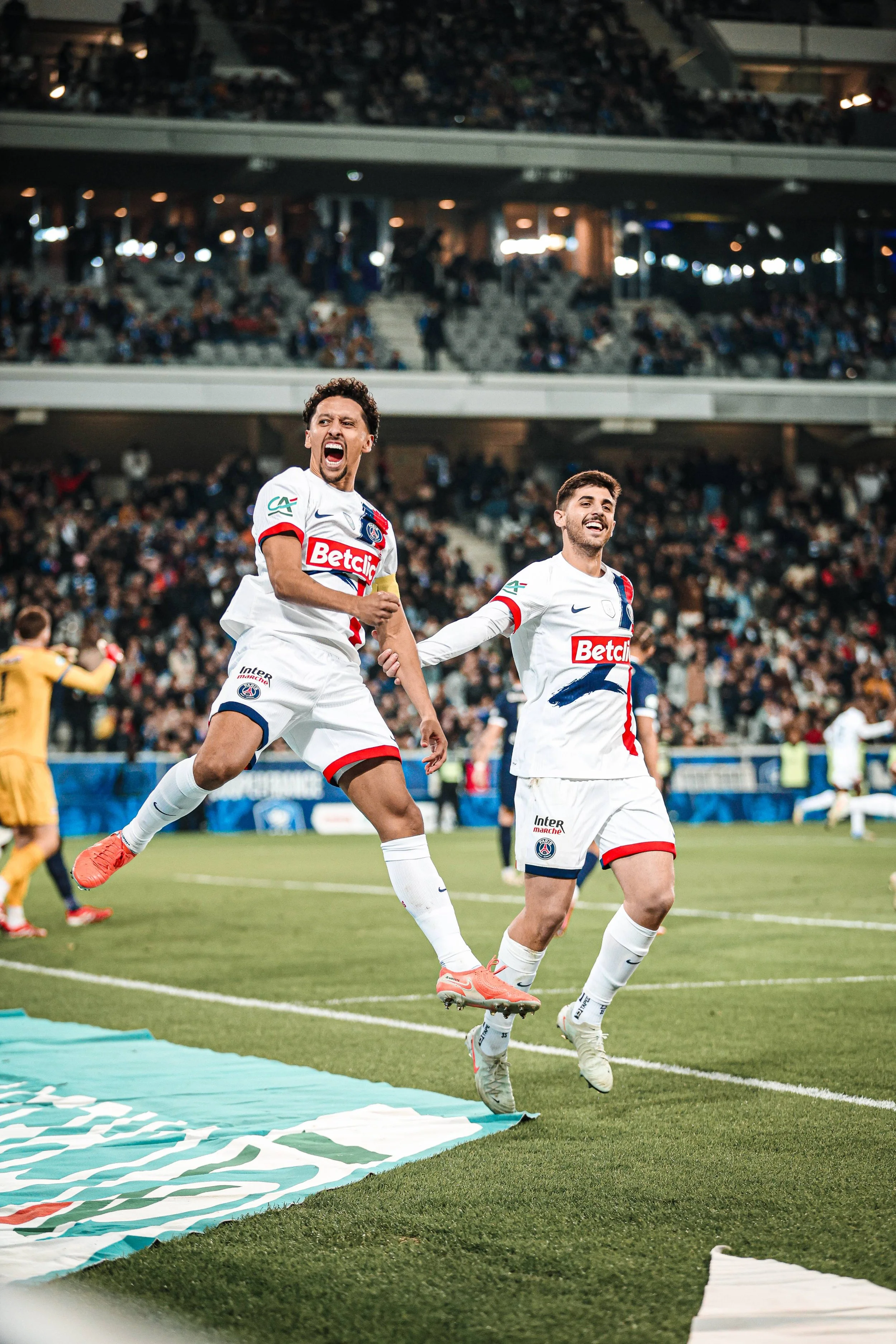 Deux joueurs de football du Paris Saint-Germain célèbrent leur victoire sur le terrain d'un stade, avec des spectateurs dans les gradins en arrière-plan.
