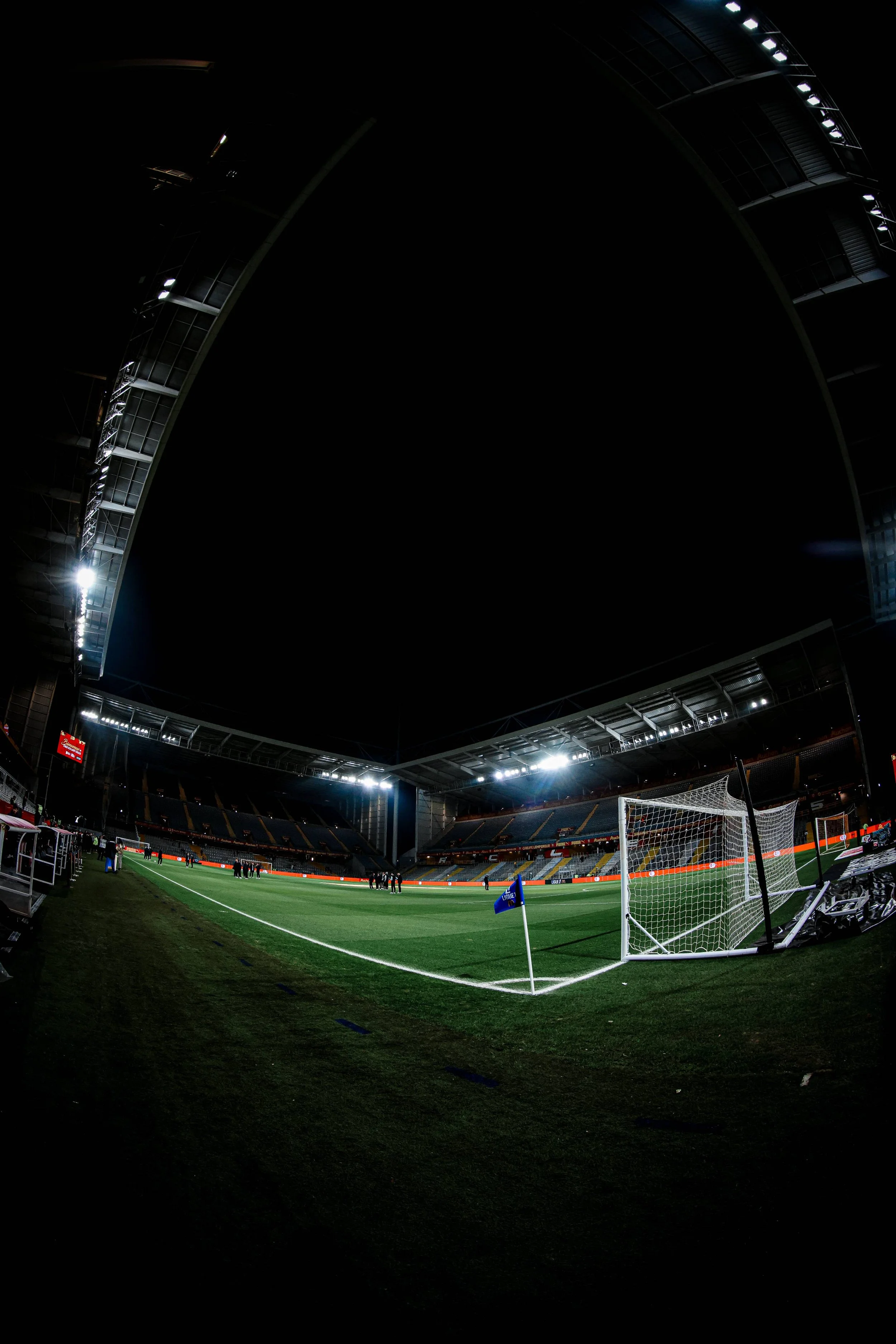Un stade de football vide avec des joueurs et des officiels au centre du terrain, vue depuis l'angle d'un but, sous un ciel nocturne.