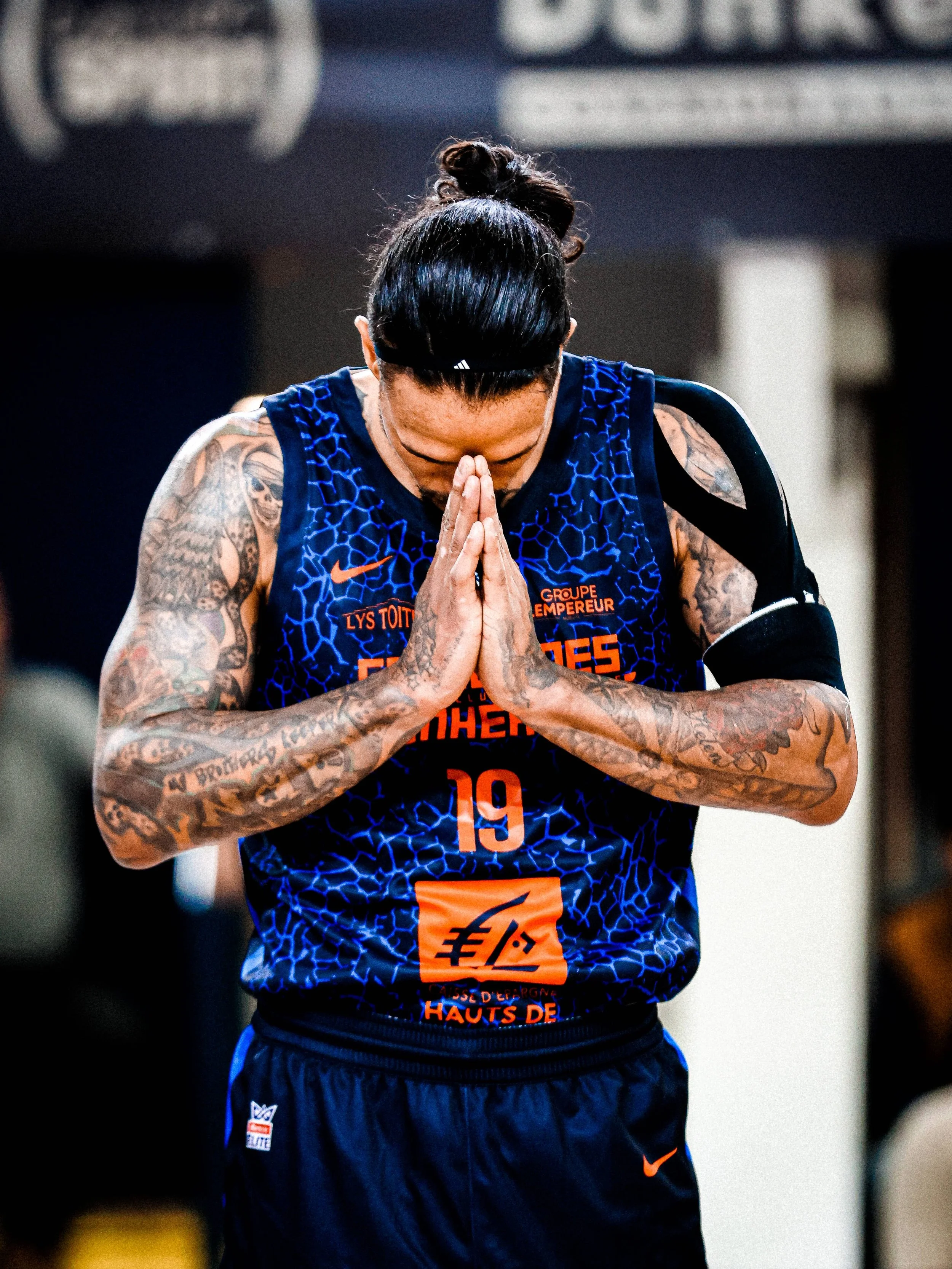 Un joueur de basket en tenue de sport, les mains jointes en priant, le regard vers le bas, avec des tatouages visibles sur les bras.