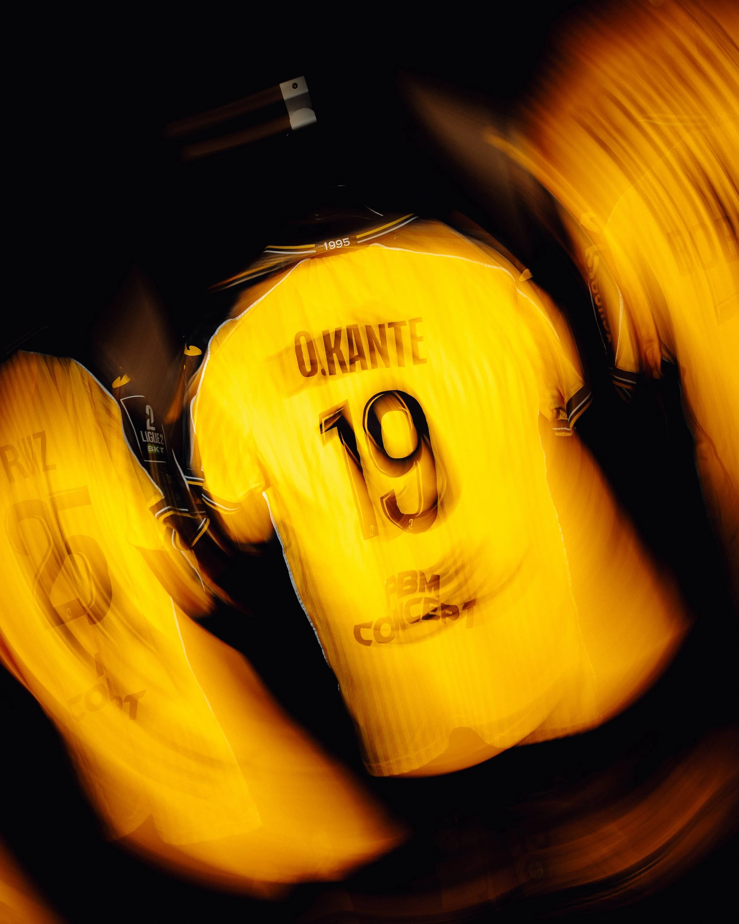 L'image montre un maillot de football jaune avec le nom 'O. KANTÉ' et le numéro '10' en noir, entouré de plusieurs maillots similaires dans un environnement sombre.