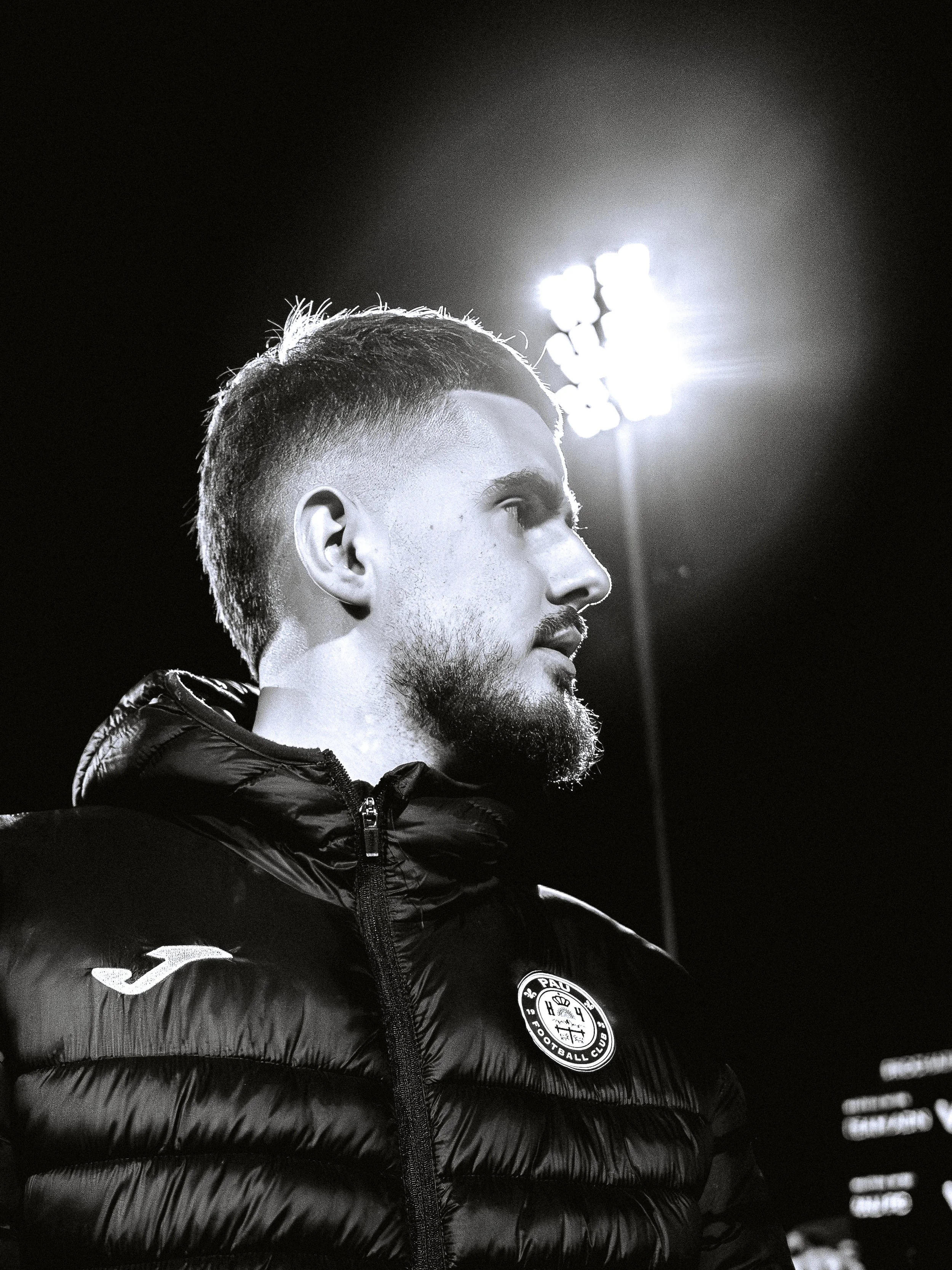 Profil d'un homme avec barbe, portant une veste de sport avec le logo du club de football PAOK, sous un éclairage intense dans un environnement sombre.