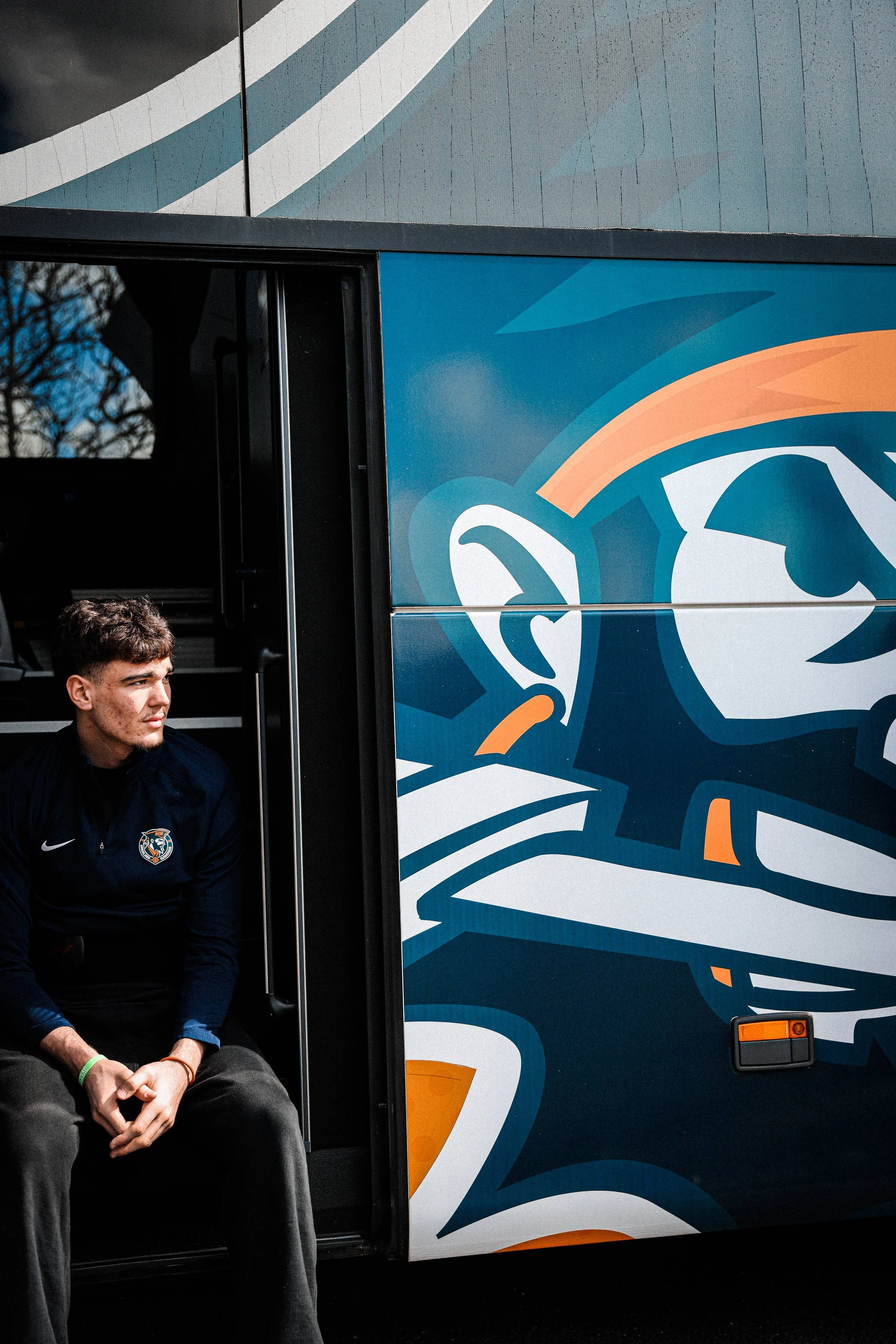 Jeune homme assis sur un bus avec une veste noire et un logo d'équipe sportive, passant devant un bus coloré avec un design de lion stylisé.