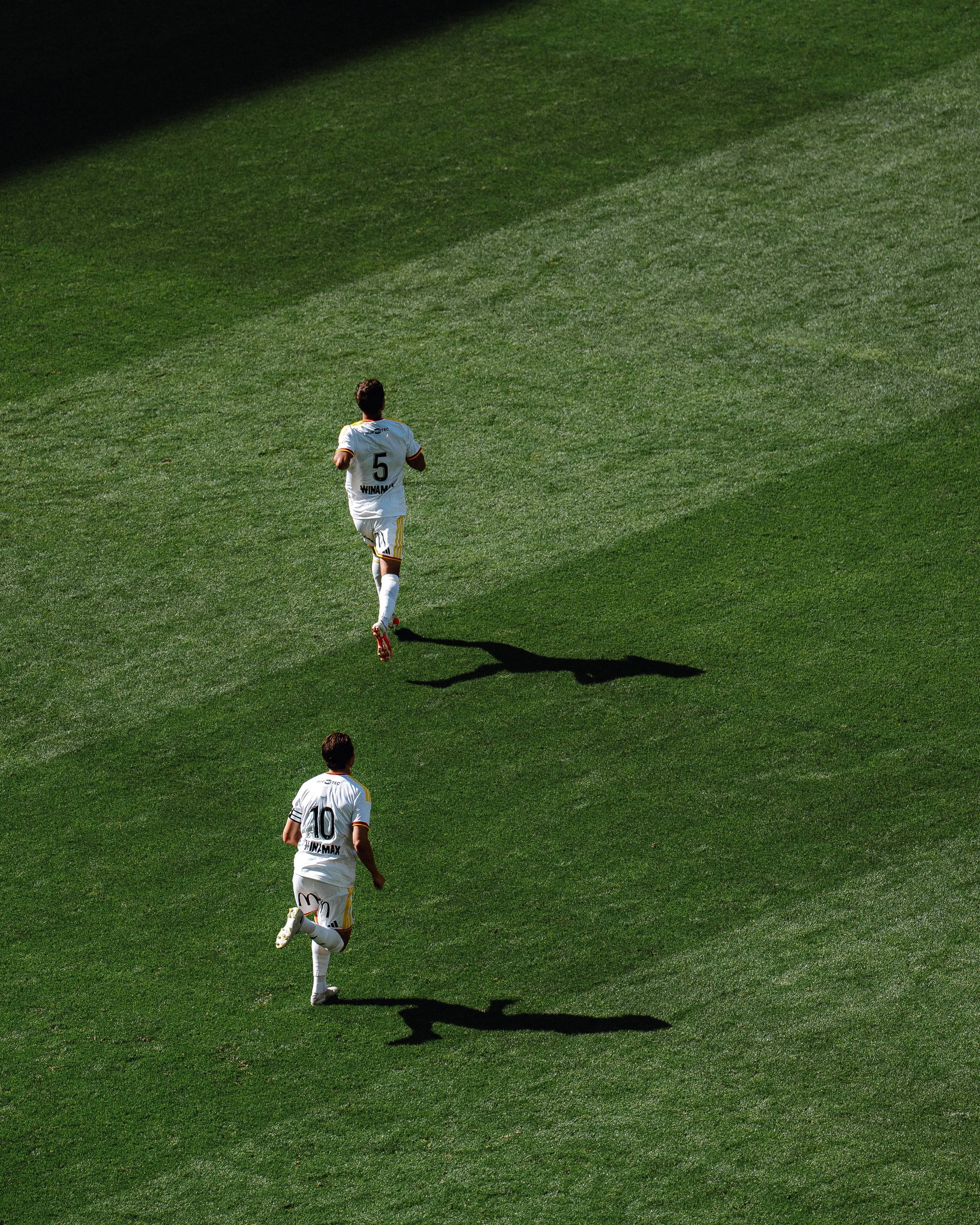 Trois joueurs de football, portant des maillots blancs avec des numéros 5, 10 et 4, courent sur un terrain en pelouse verte lors d'un match.