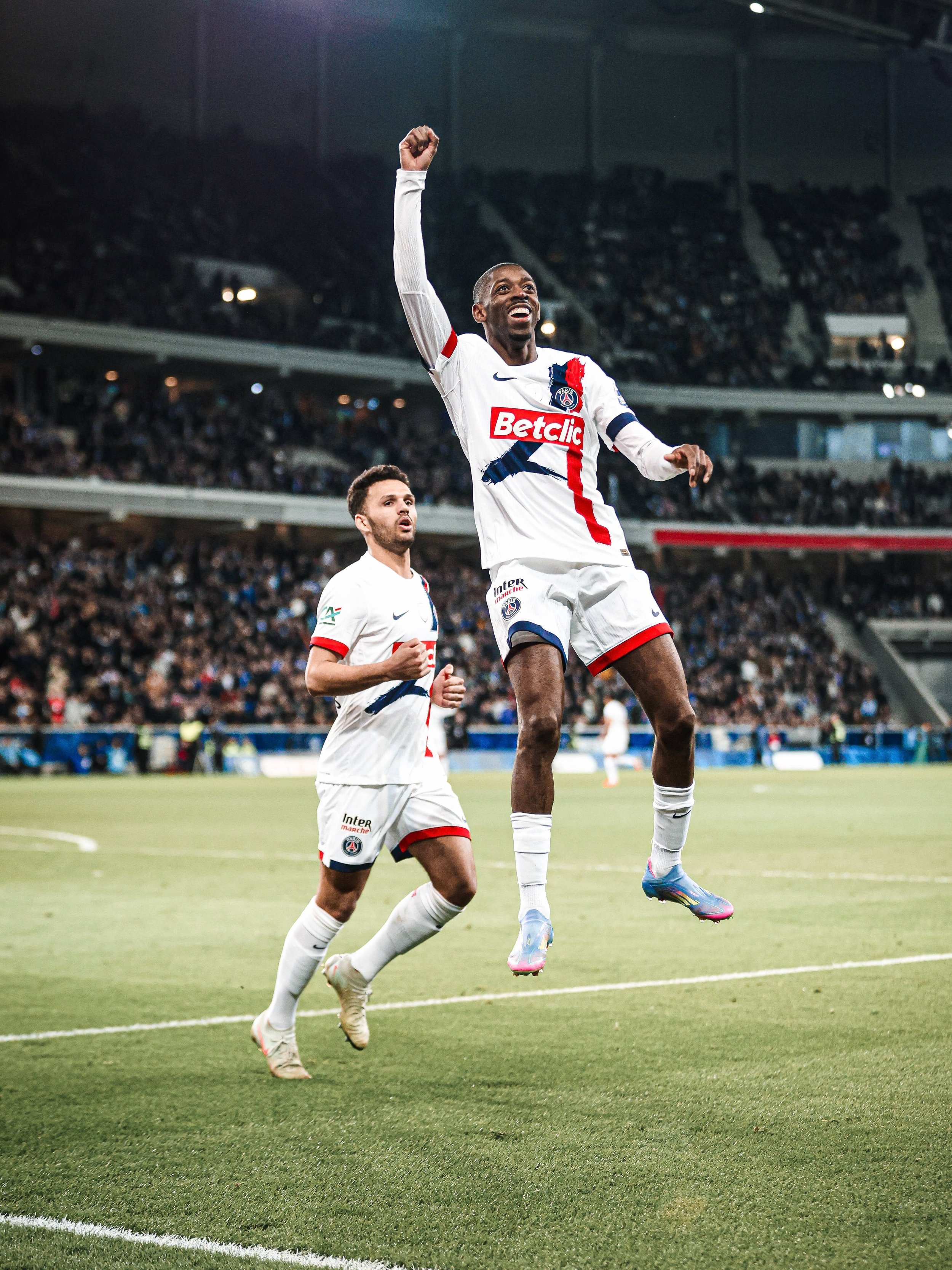 Deux joueurs de football du Paris Saint-Germain célèbrent lors d'un match, l'un sauteur dans les airs avec un bras levé, l'autre courant derrière lui, dans un stade rempli de spectateurs.