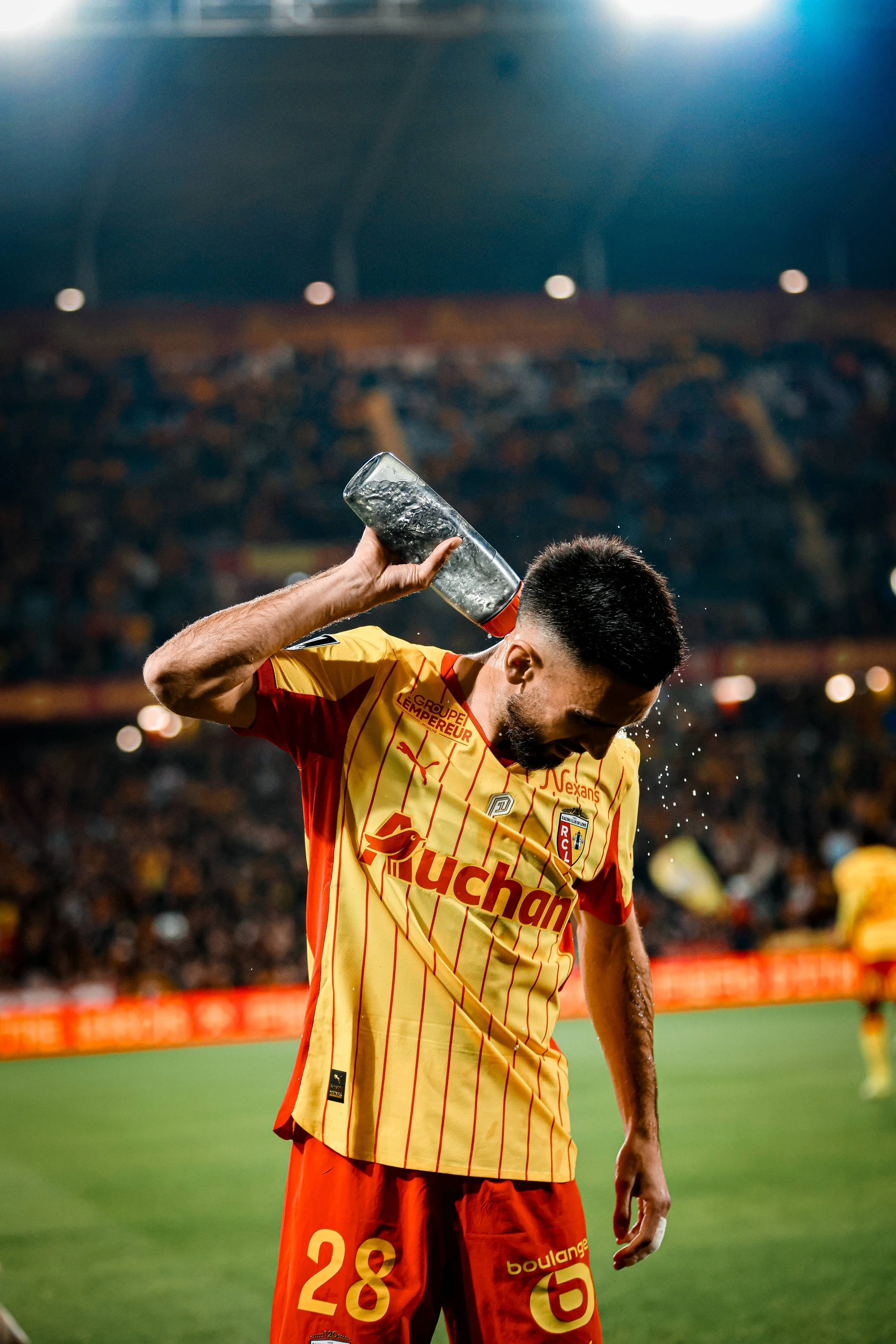 Un joueur de football en tenue jaune et orange, portant le numéro 28, verse de l'eau sur sa tête lors d'un match, avec un public dans les gradins en arrière-plan.