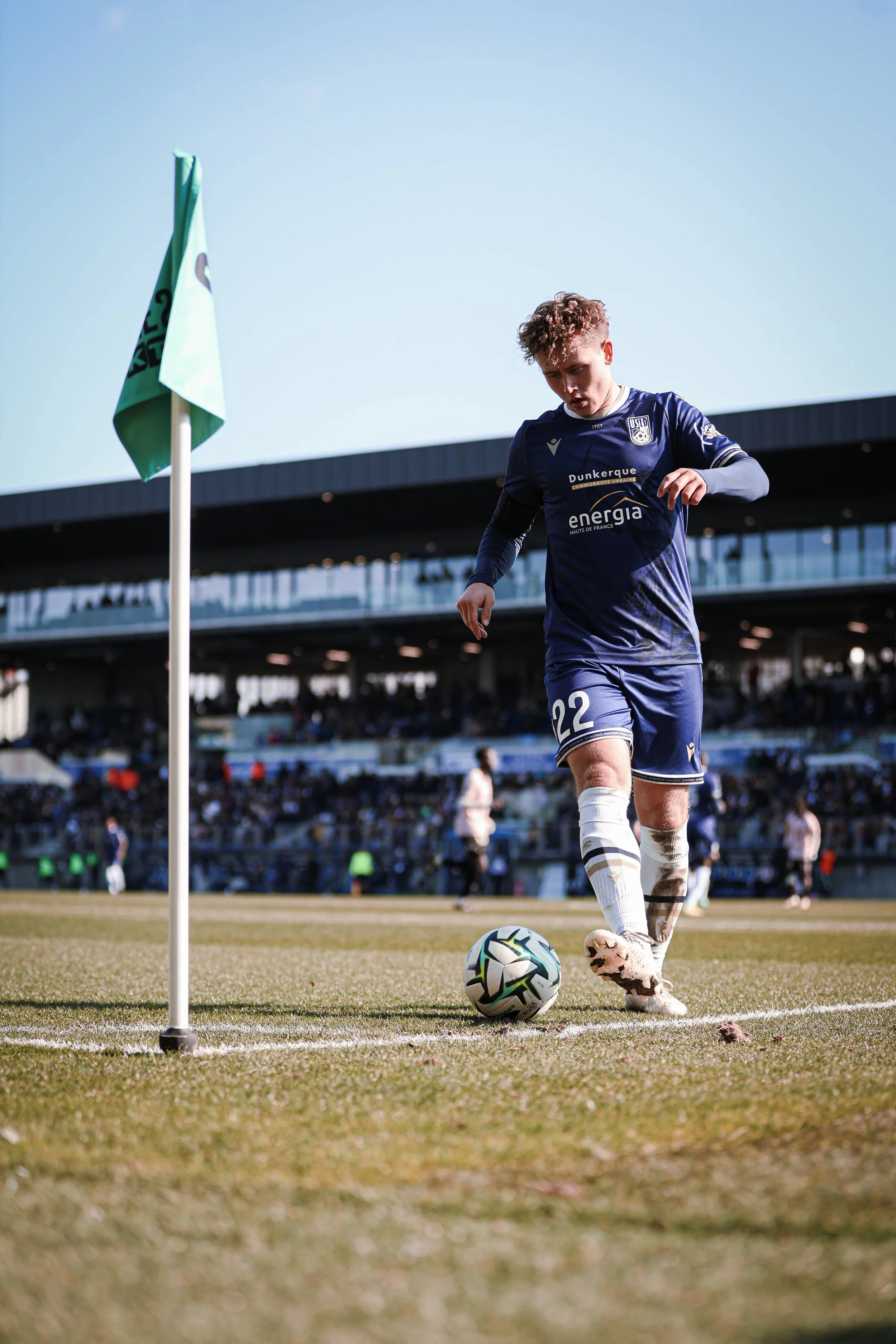 Un joueur de football en uniforme bleu, portant le numéro 22, se prépare à tirer un coup de pied lors d'un match sur un terrain herbeux, près d'une pièce de drapeau, dans un stade rempli de spectateurs.
