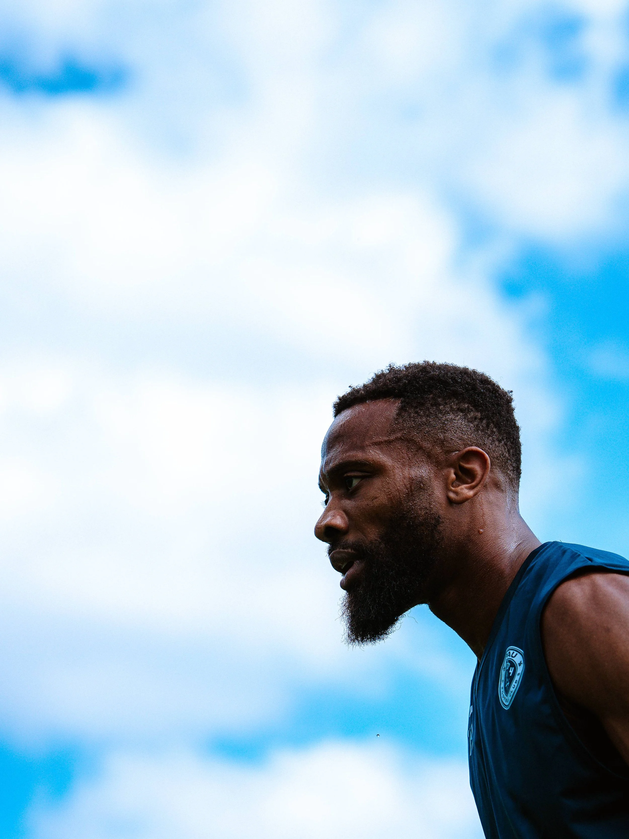 Un homme noir, probablement un athlète, en vêtements de sport bleus, vu de profil, sous un ciel bleu clair avec quelques nuages.