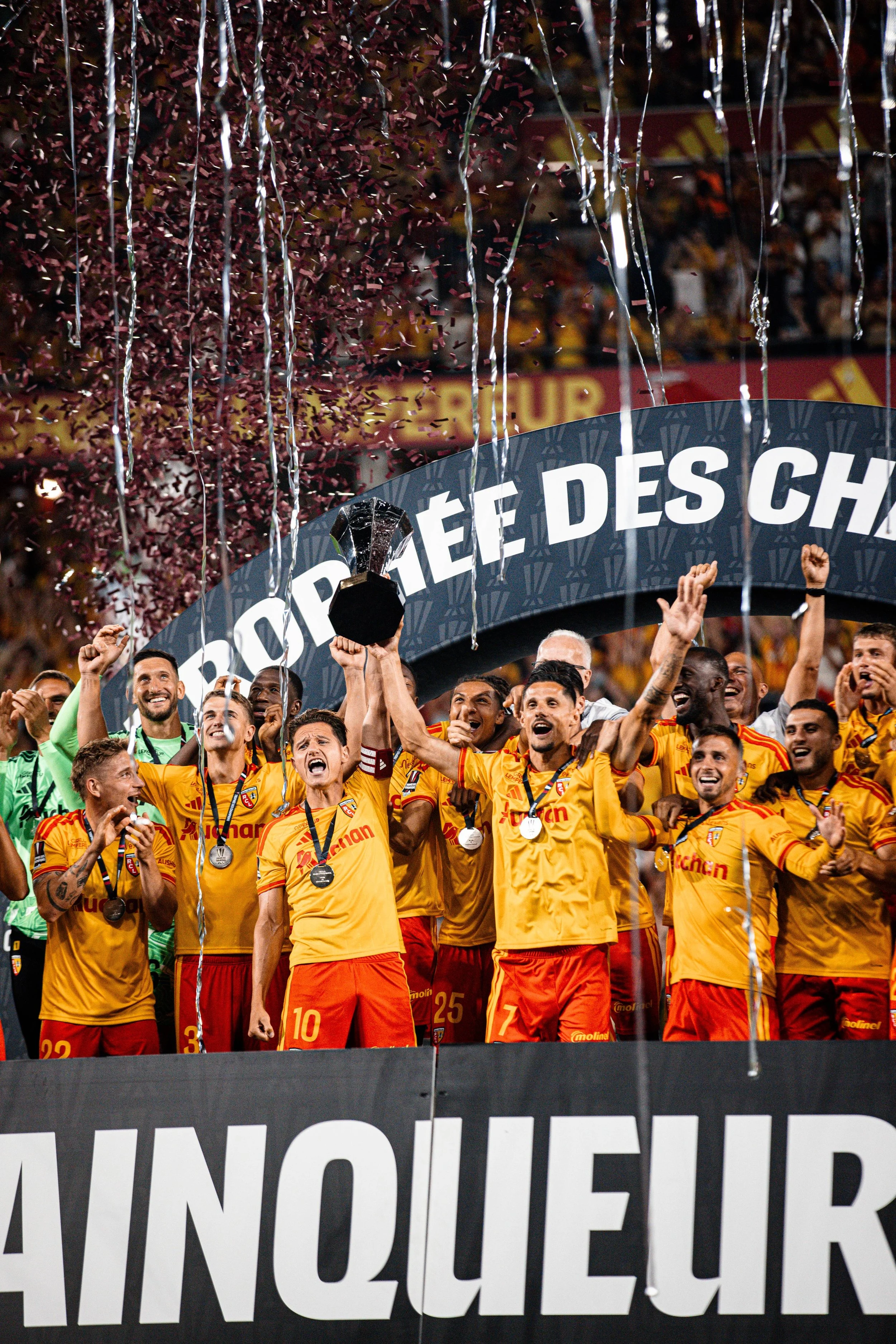 Une équipe de football portant des maillots jaunes et rouges célèbre sur un podium, tenant un trophée à la main, avec des confettis et des rubans argentés, lors d'une cérémonie de remise de prix.