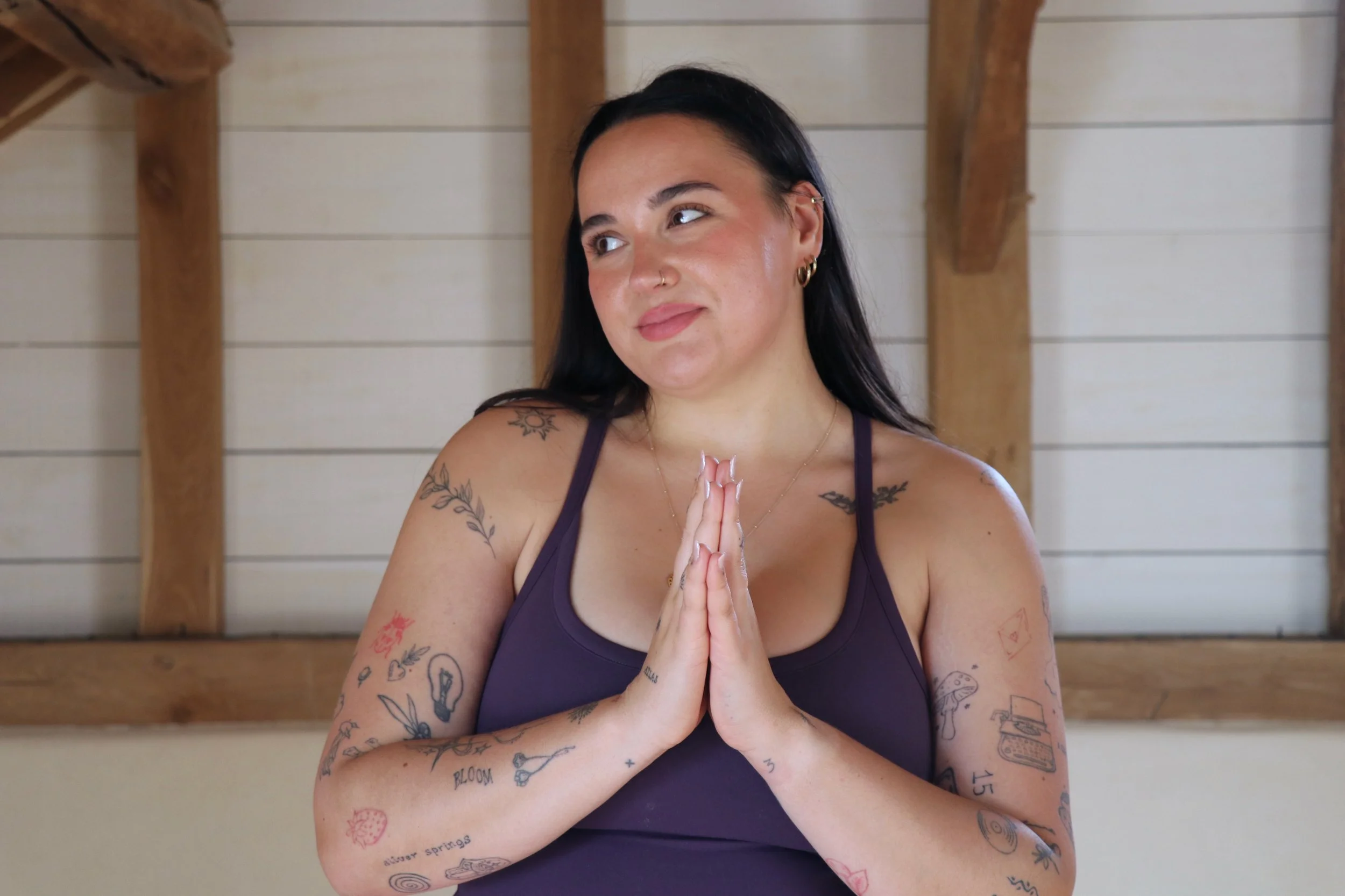 A woman with black hair and tattoos on her arms and shoulders is standing indoors with hands in a prayer position. She is wearing a purple tank top and has a peaceful expression.