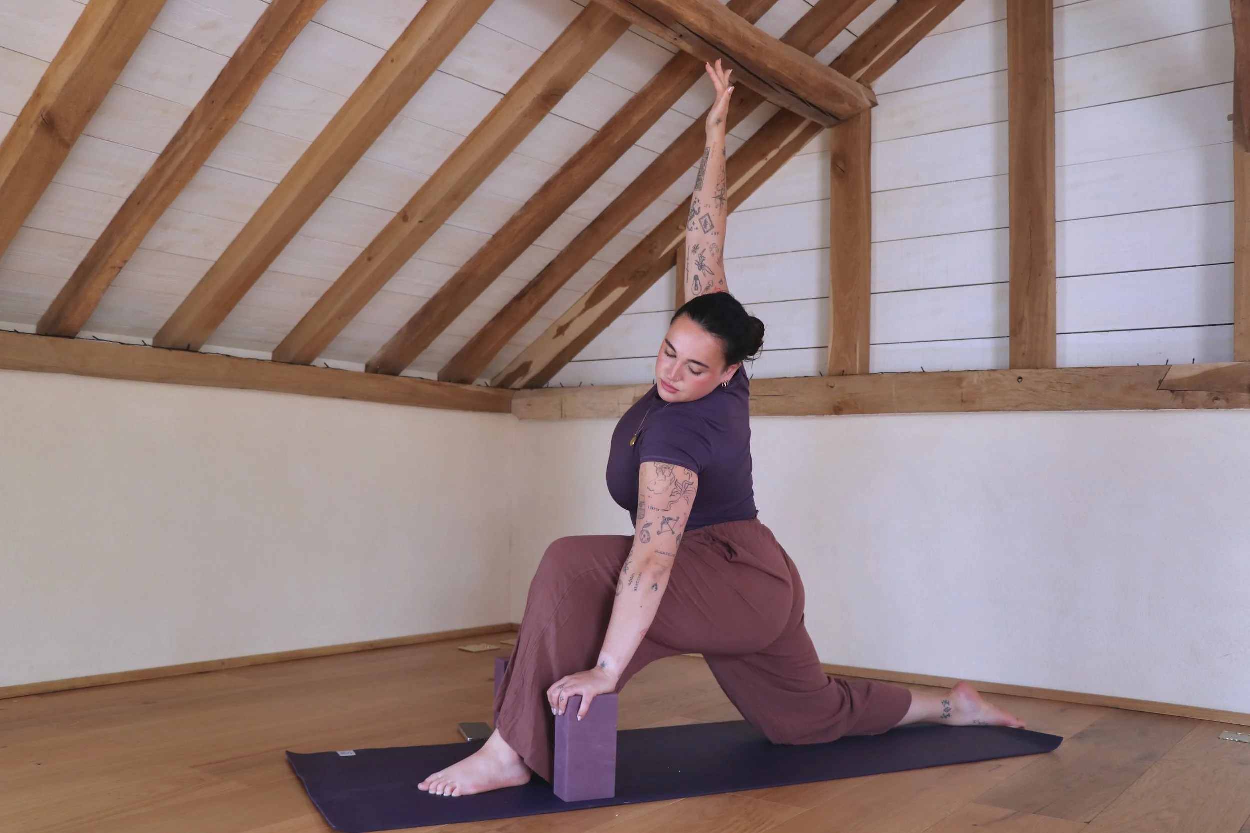 A woman practicing yoga indoors, stretching in a low lunge position with one arm extended upward and the other arm reaching down. She is on a yoga mat in a room with wooden floors and a wooden ceiling with exposed beams.