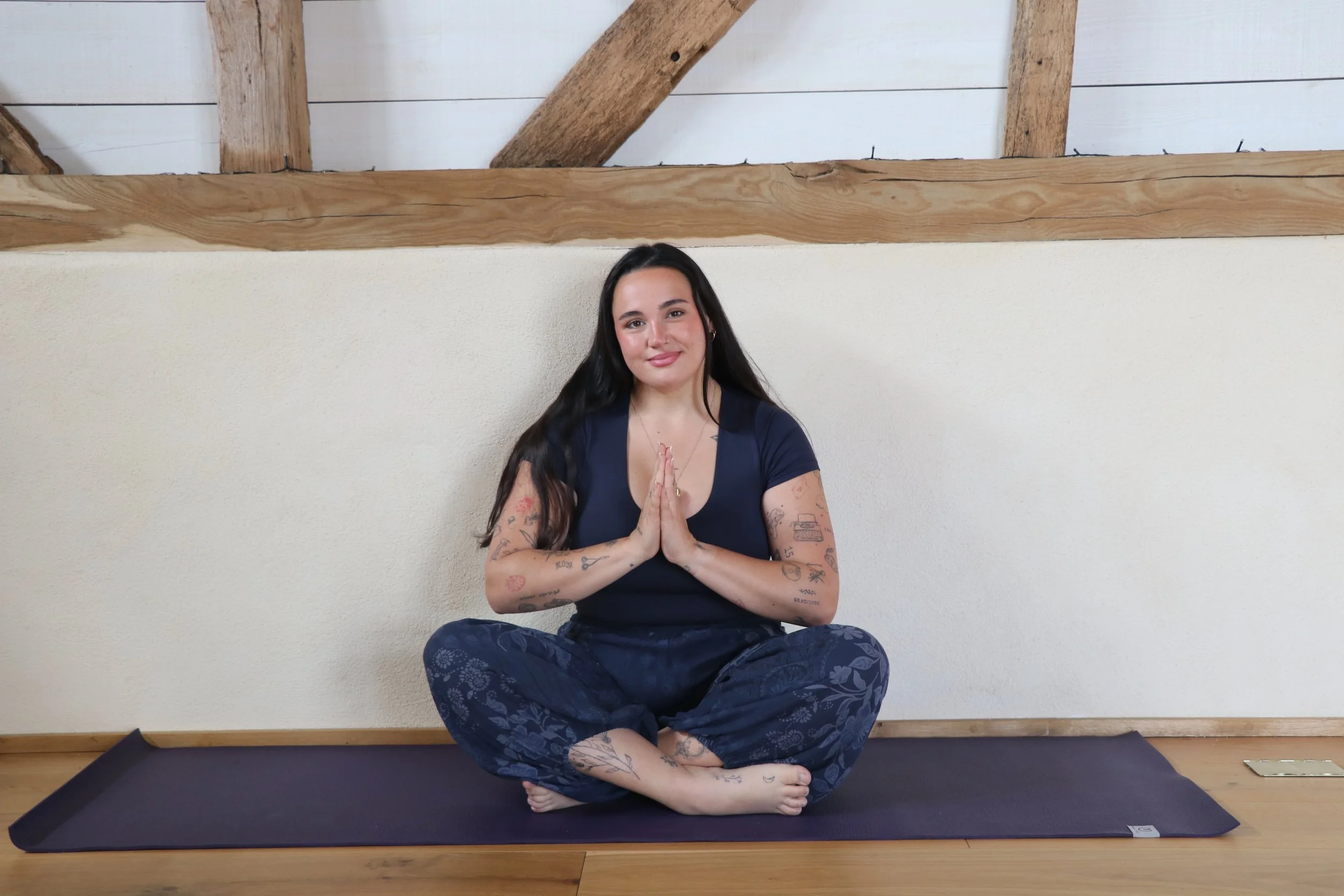 Woman with long dark hair, tattoos, wearing a dark blue top and patterned pants, sitting cross-legged on a yoga mat indoors, practicing meditation with hands pressed together in a prayer position, smiling gently, against a white wall with wooden beams.