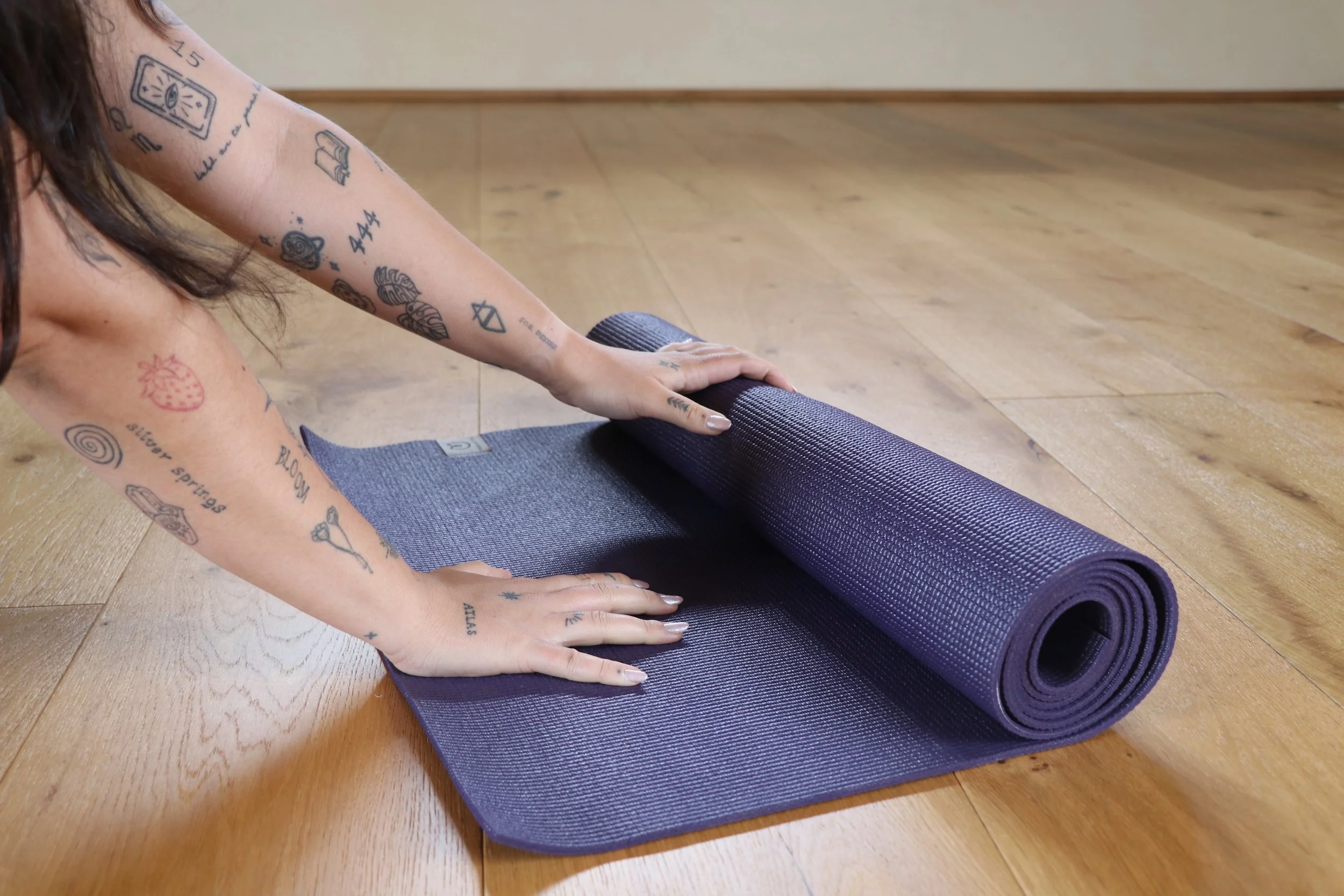 A person with tattoos on their arms, wearing a sleeveless top, is placing a purple yoga mat on a wooden floor in a room with plain walls, preparing for a yoga or exercise session.