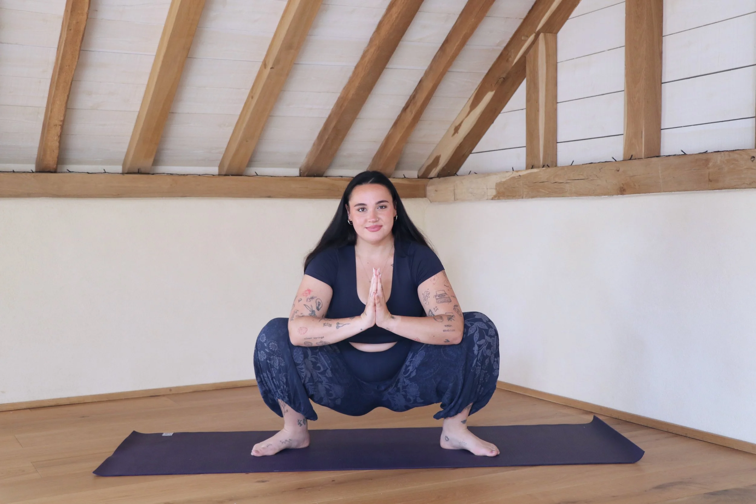 A woman practicing yoga indoors, squatting on a mat with hands in prayer position, in front of a wall with exposed wooden beams on the ceiling.