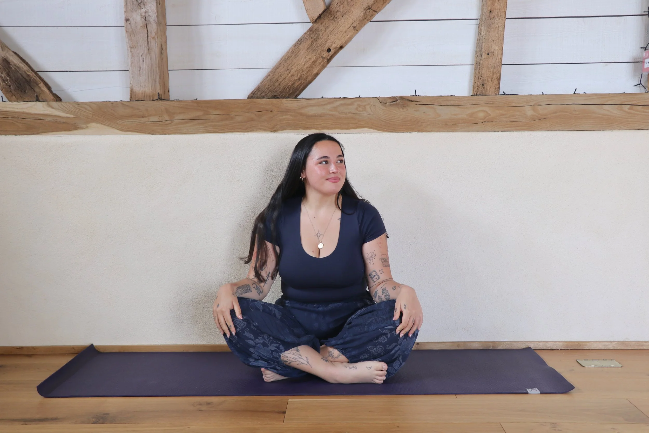 A woman with tattoos on her arms and legs sits cross-legged on a yoga mat, meditating in front of a white wall with exposed wooden beams on the ceiling.