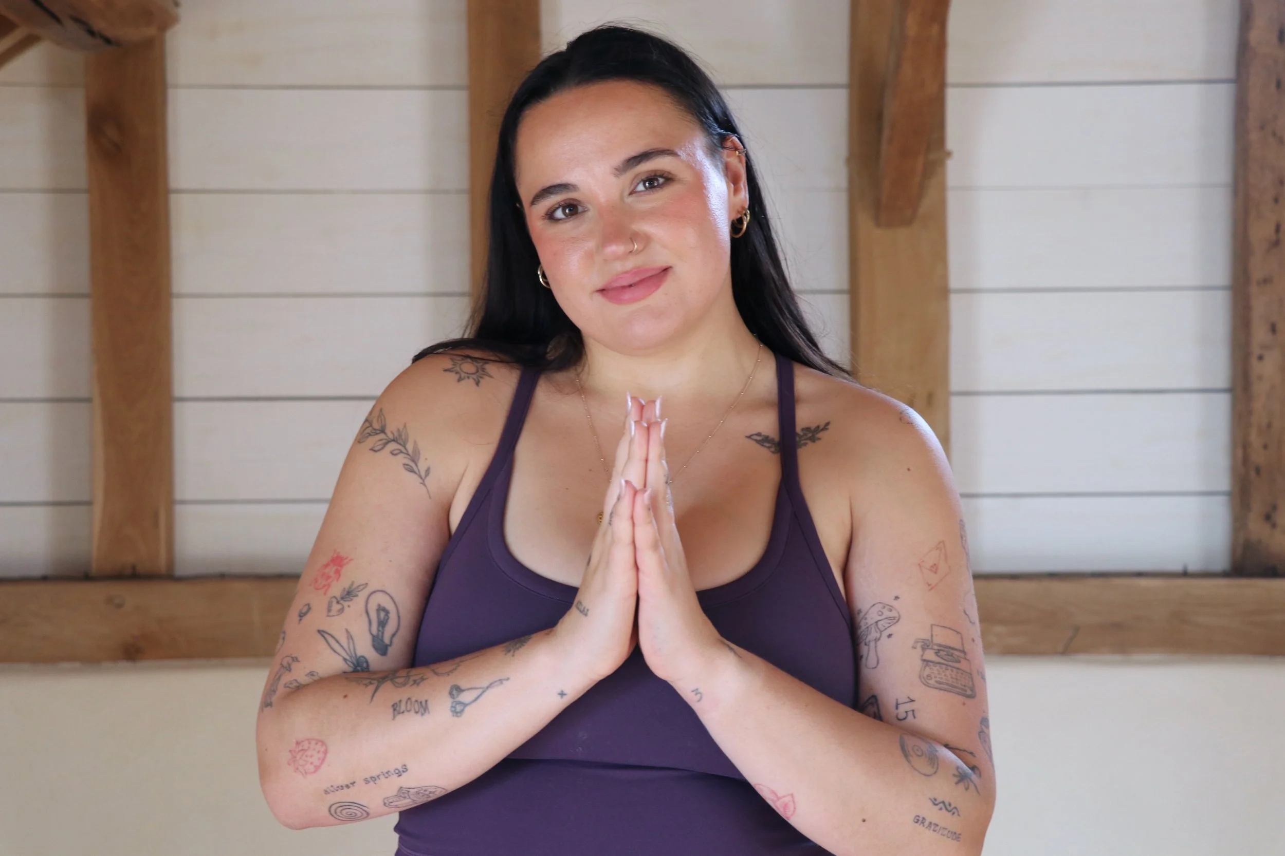 A woman with long dark hair and tattoos on her arms and shoulders, wearing a purple tank top, standing with her hands pressed together in a prayer pose, indoors with wooden beams and shiplap walls behind her.