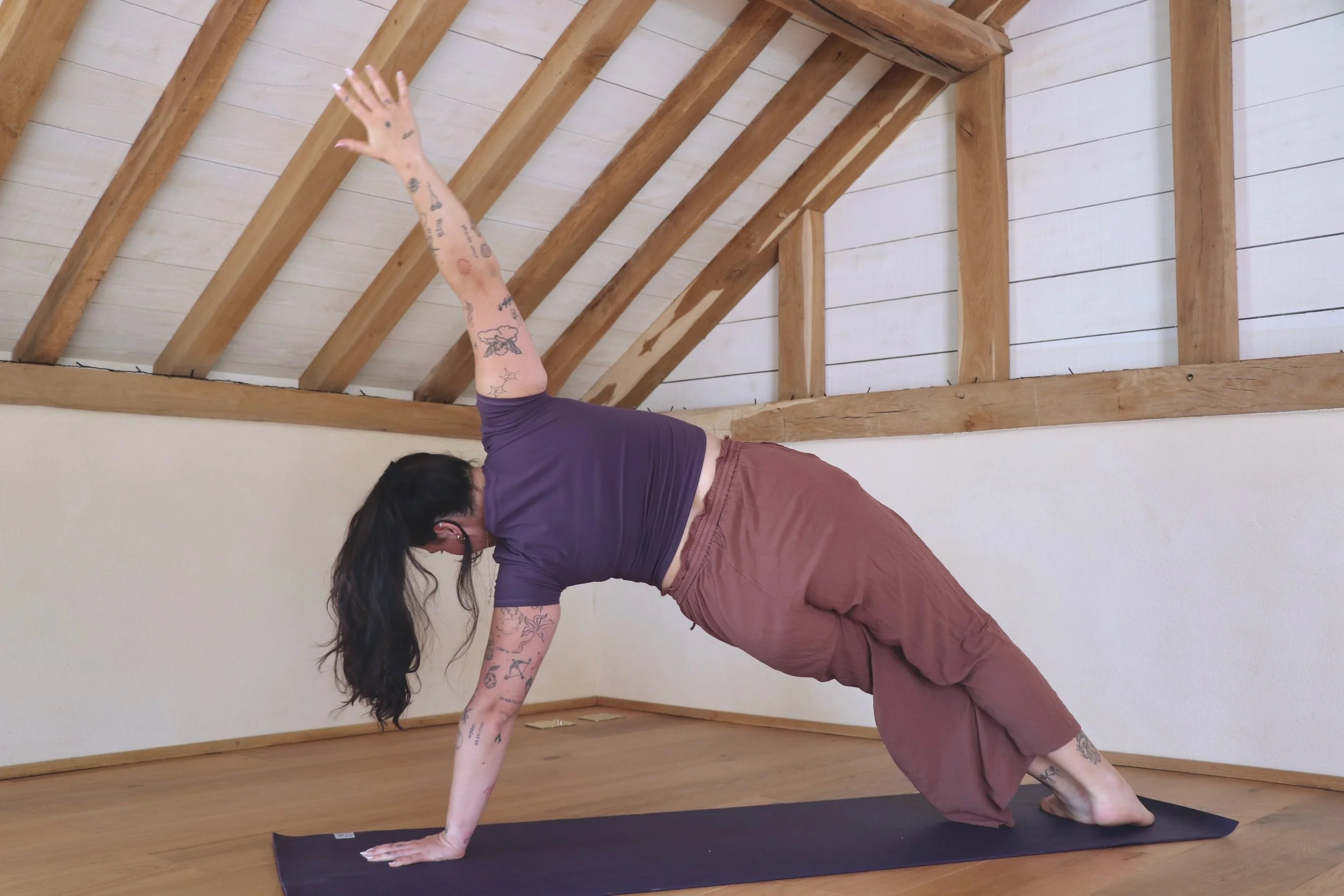A woman practicing yoga indoors on a black yoga mat in a room with wooden beams and white walls, wearing a purple top and maroon pants.