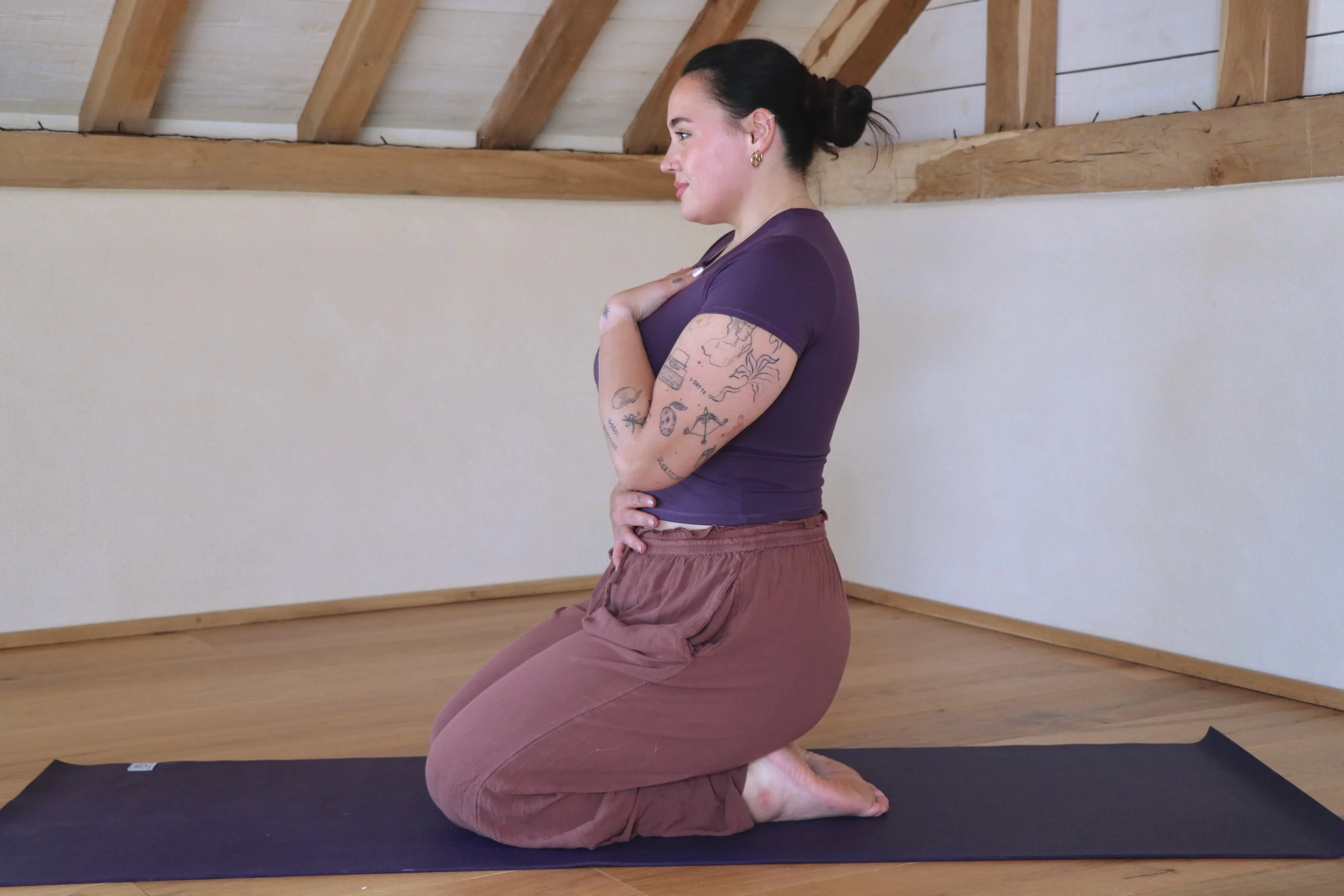 A woman practicing yoga indoors on a black mat, kneeling with her hands on her chest, wearing a purple top and brown pants, in a room with wooden beams on the ceiling and light-colored walls.