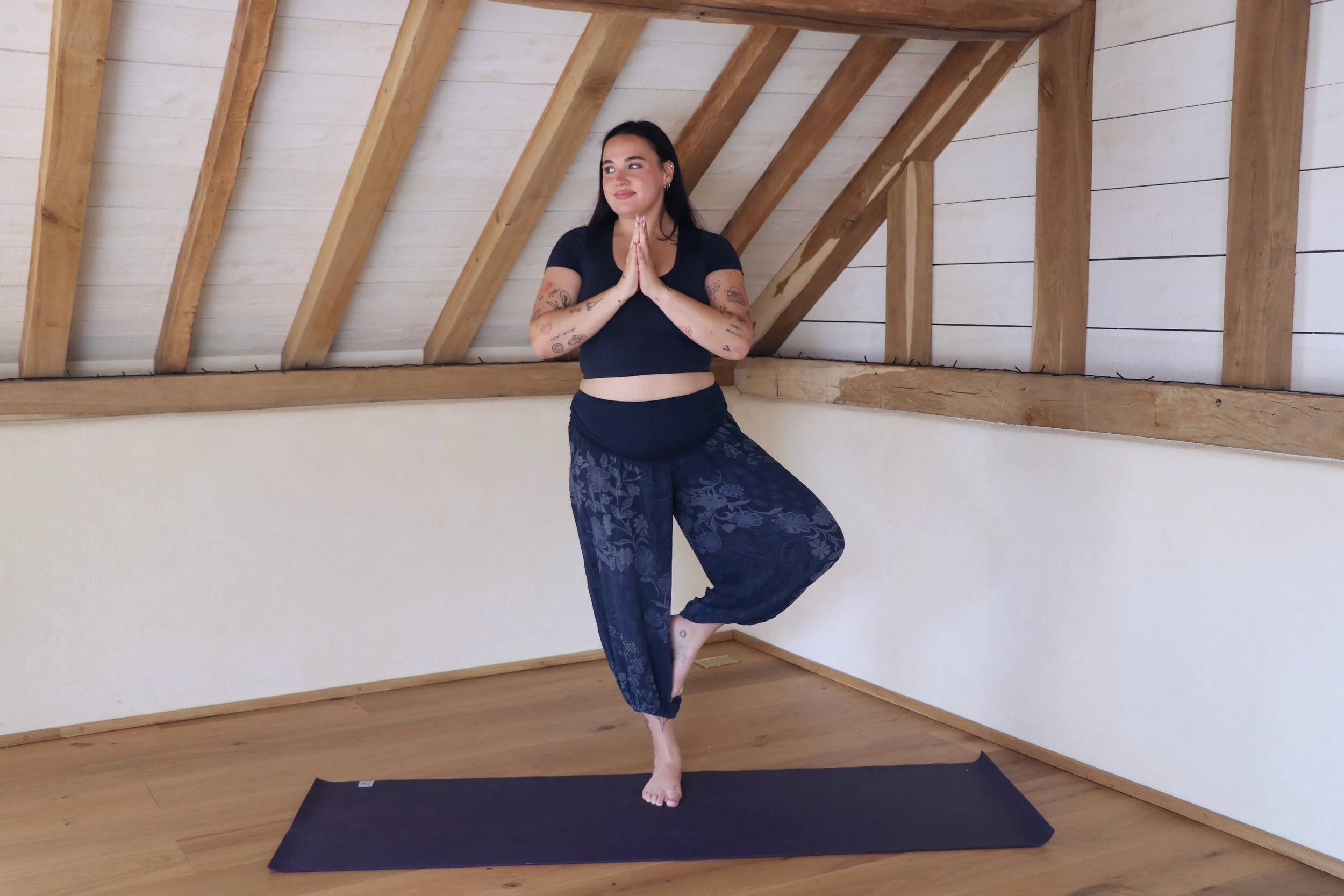 A woman with dark hair, tattoos on her arms, in a black shirt and patterned pants, practicing yoga in a room with wooden beams and white walls, standing on a black yoga mat in a tree pose with palms pressed together.