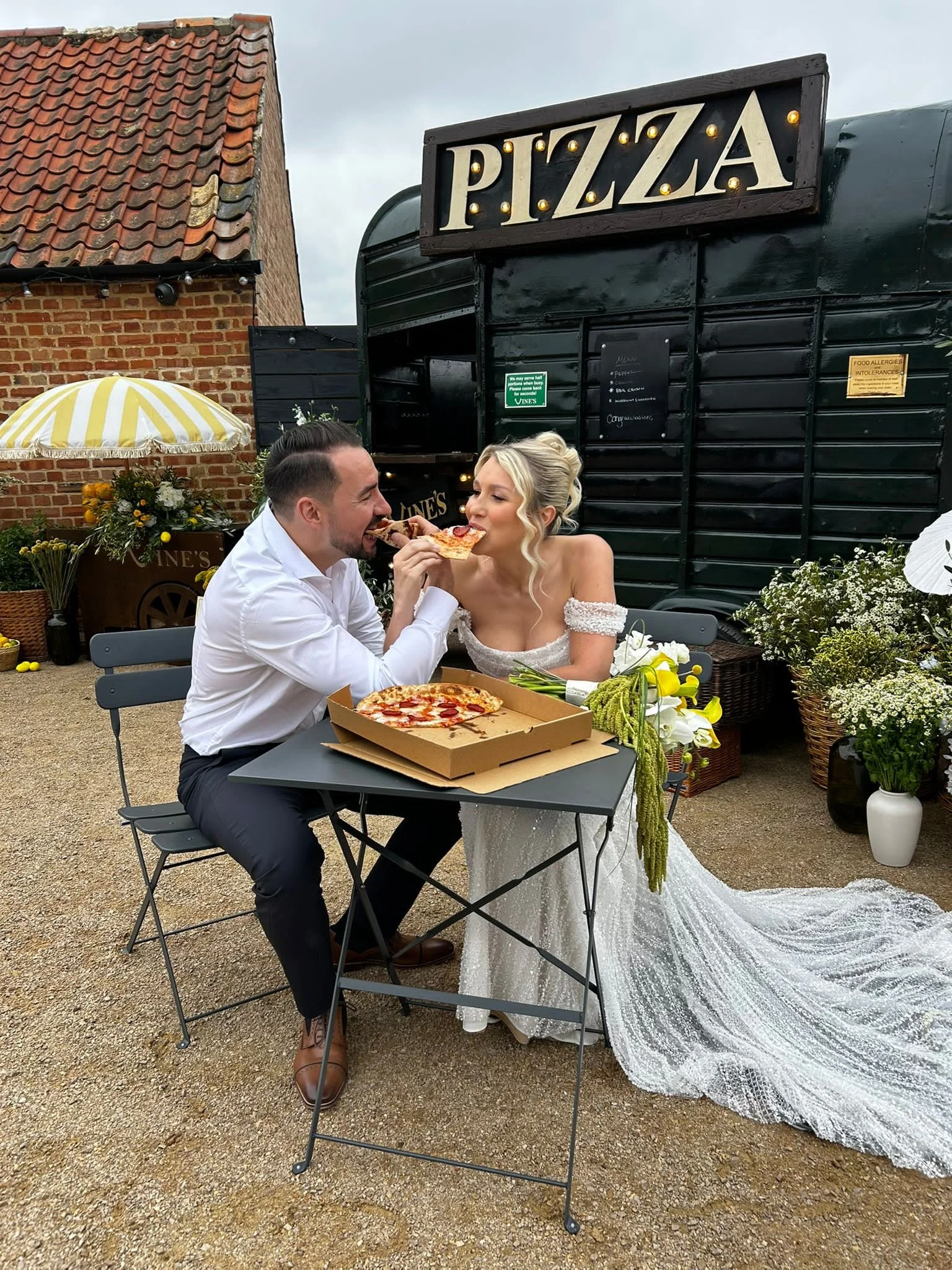 A man and woman in wedding attire sharing a slice of pizza at an outdoor celebration, with a pizza box on the table and wedding flowers nearby.