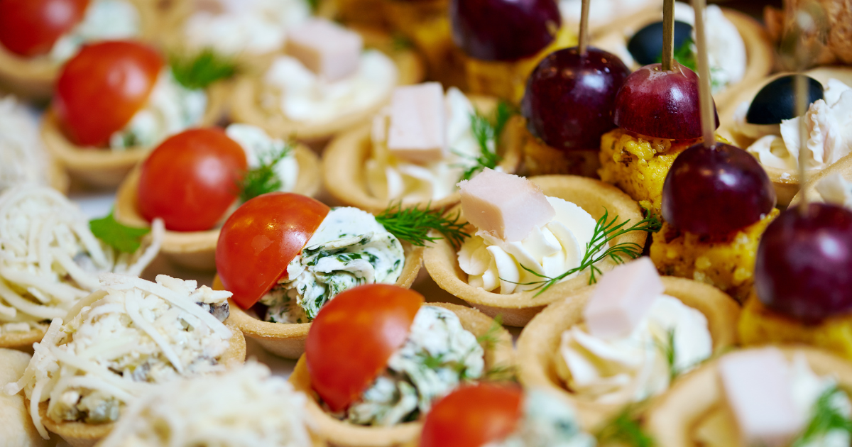 Assorted bite-sized savory appetizers on a platter, including cherry tomato and herb pastries, cheese and ham tartlets, and mini skewers with grapes, pineapple, and meat or cheese.