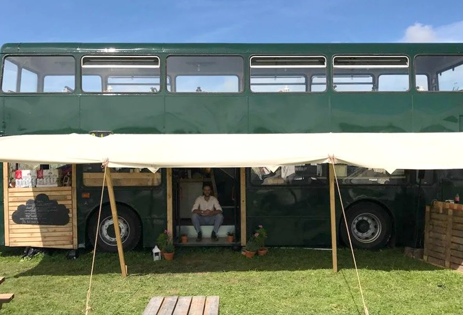 Green double-decker bus converted into a cafe with outdoor seating area, under a canopy, on a grassy field.