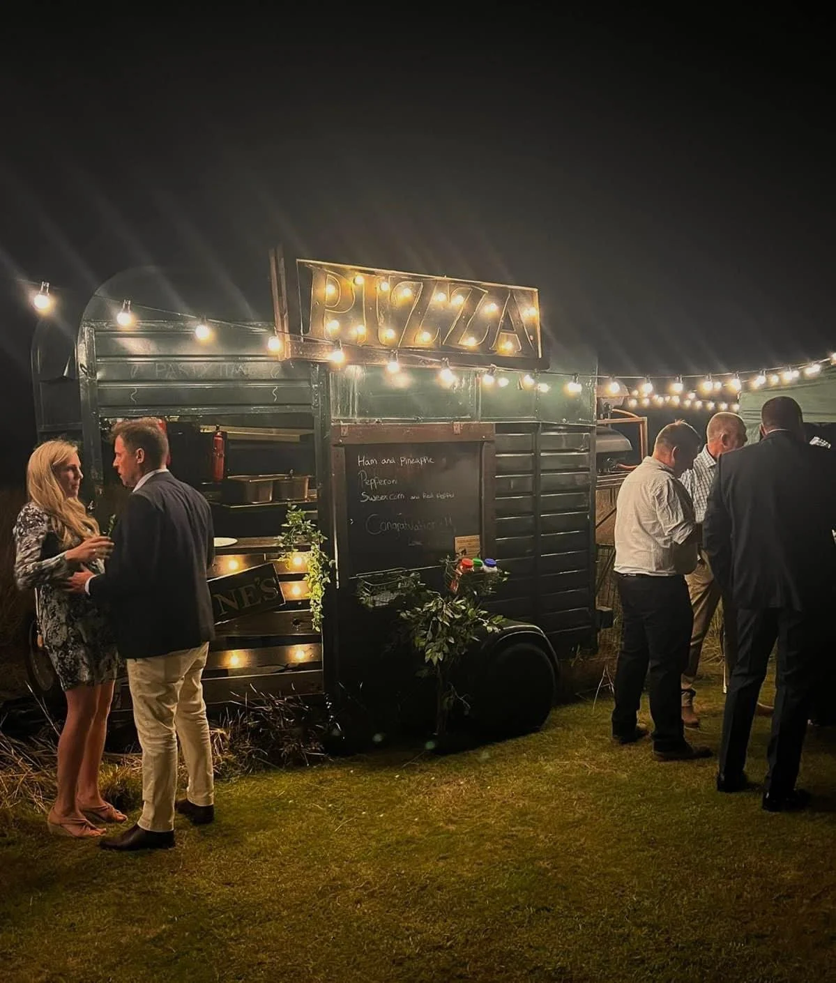 Outdoor pizza food truck with people gathered at night, illuminated by string lights, menu board displaying options, and decorations with plants.