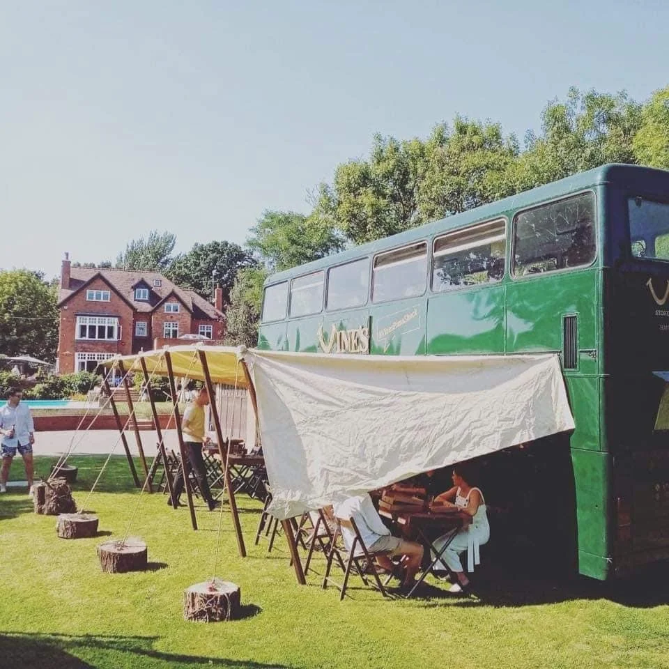 Green double-decker bus converted into a café with outdoor tent seating, people sitting under the tent, grassy area, and a brick house in the background.