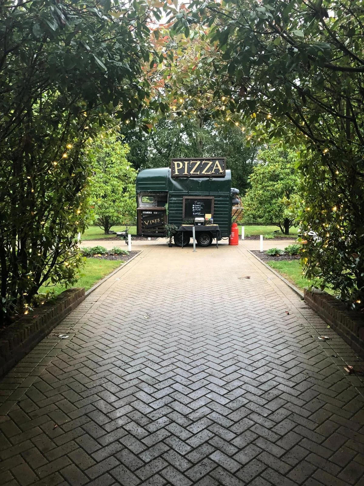Pizza truck on a pathway surrounded by trees with string lights at dusk.