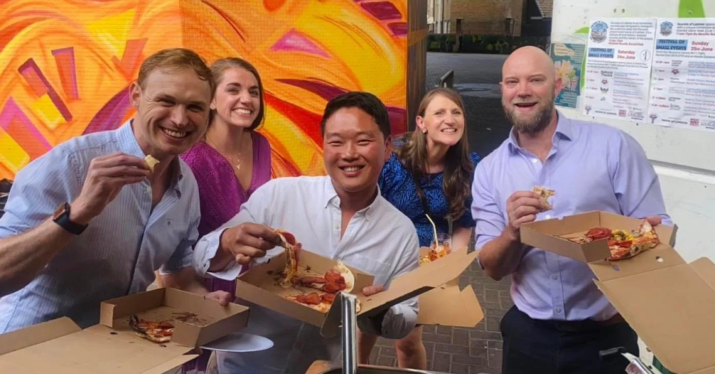 Group of people holding pizza boxes and slices, smiling outdoors with colorful mural background.