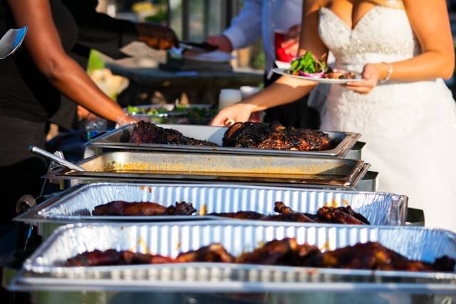 Buffet table with trays of food at an outdoor event, people serving themselves, one wearing a wedding dress.