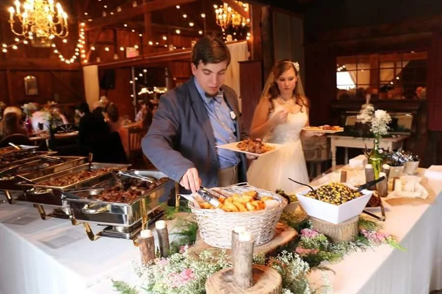 Guests at a wedding buffet selecting food from a table decorated with flowers and candles in a rustic venue with string lights.