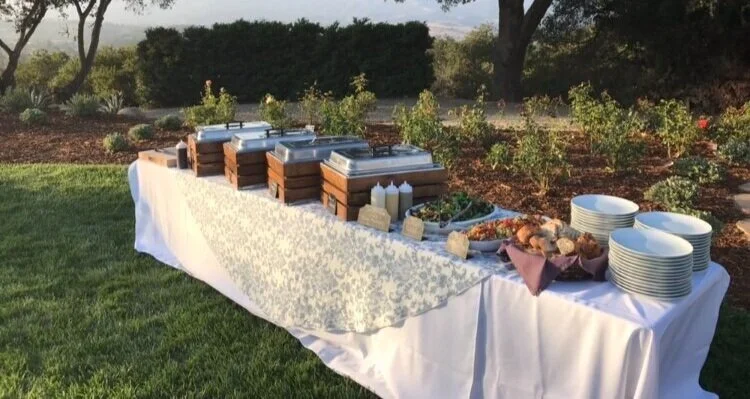 Outdoor buffet table setup with chafing dishes and serving platters in a garden setting.