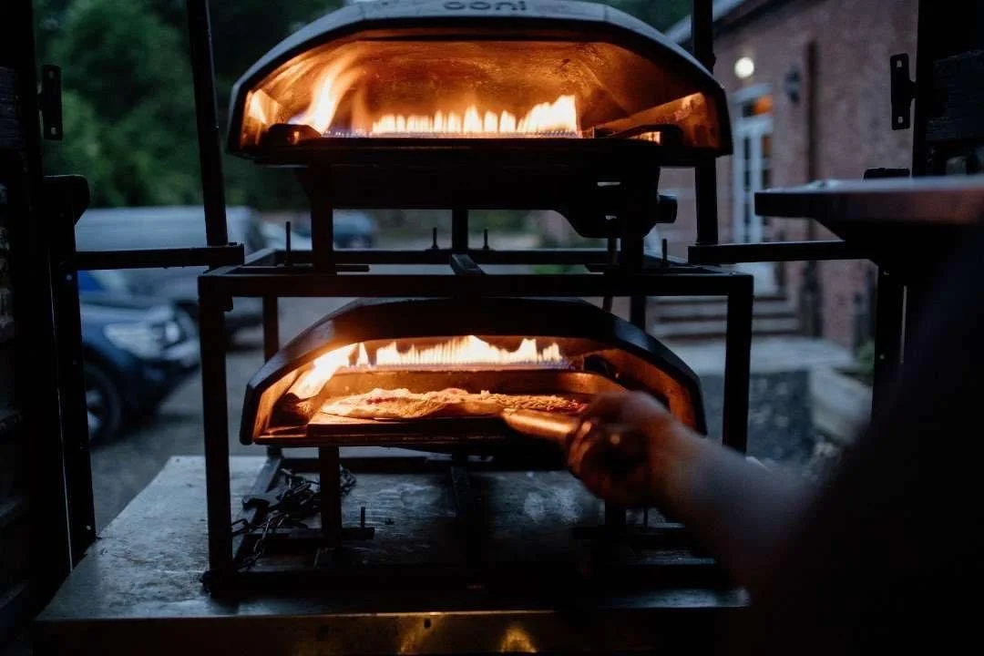 Outdoor pizza oven with flames, pizza being inserted, evening light.