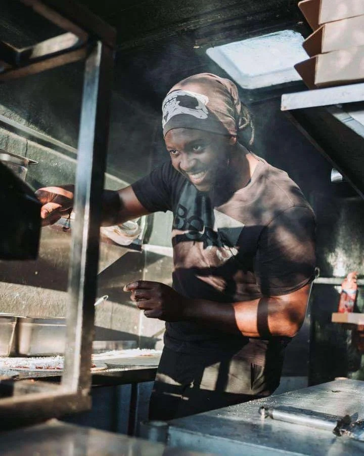 Person cooking inside a food truck, wearing a headscarf and black T-shirt, smiling while working at the grill in a well-lit kitchen.
