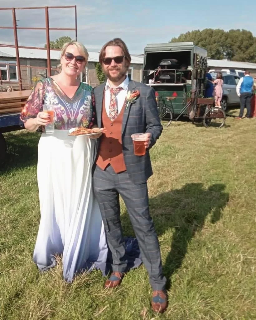 A man in a suit and a woman in a colorful top and white skirt stand on grass, holding drinks, with a food truck in the background at an outdoor event.
