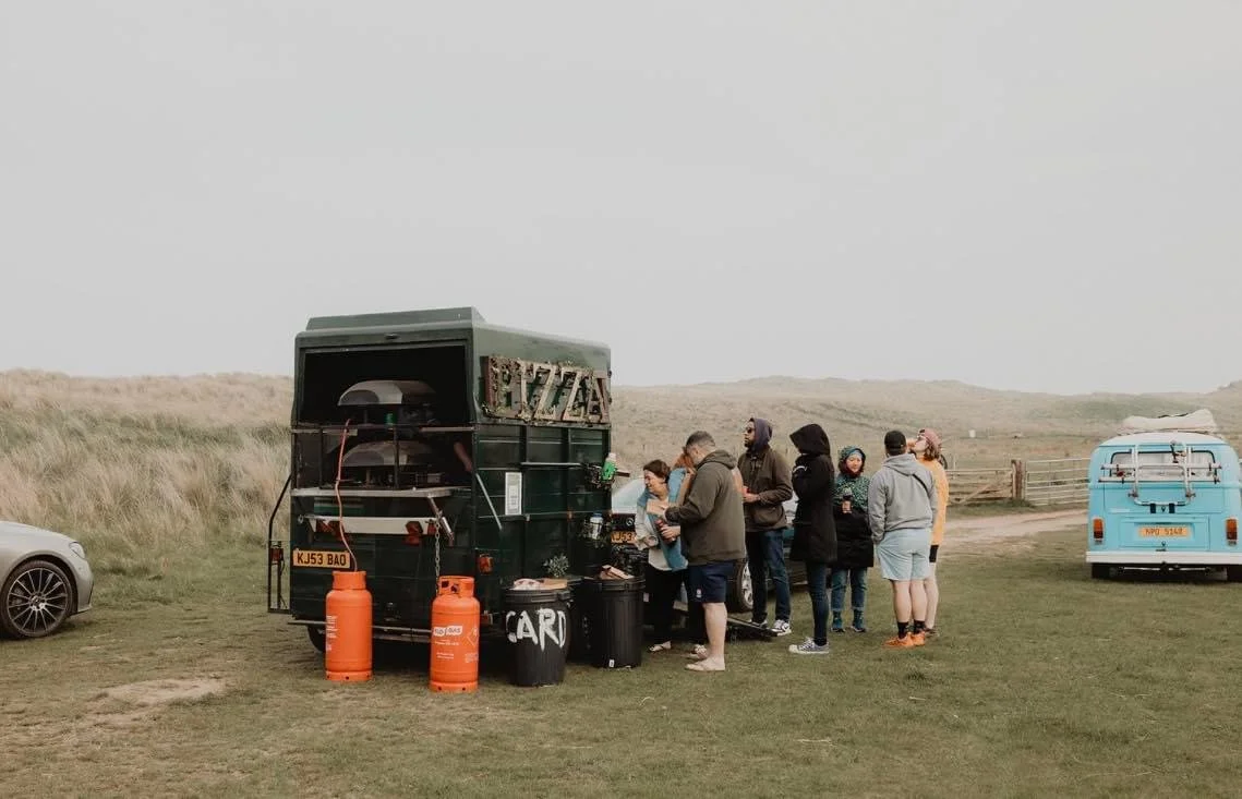 People standing in line at a green pizza truck with two orange gas cylinders and a blue van nearby, set in a grassy outdoor area with hills in the background.