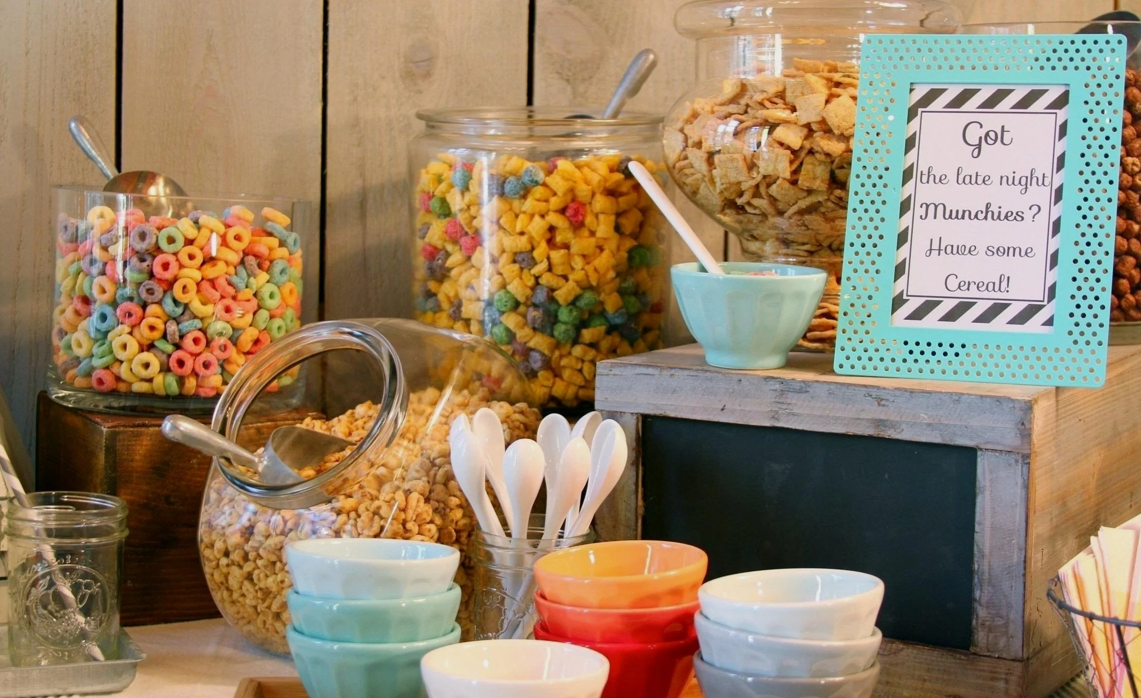 A variety of cereals in glass jars with colorful bowls, spoons, and a sign promoting late-night cereal munchies.