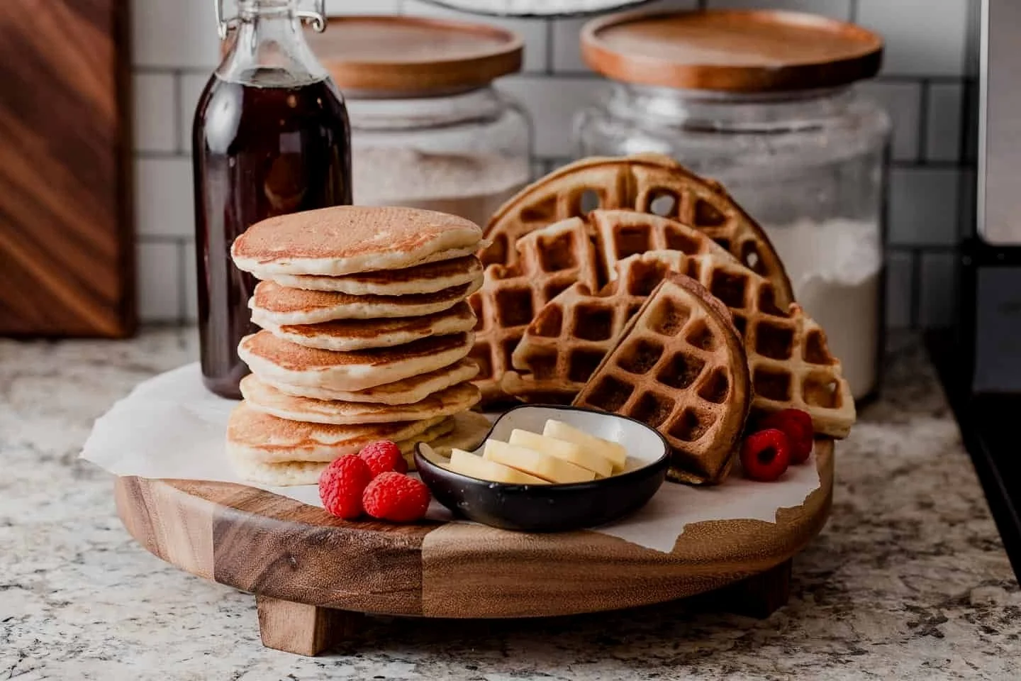 Breakfast platter with pancakes, waffles, butter slices, raspberries, and syrup on a wooden tray.