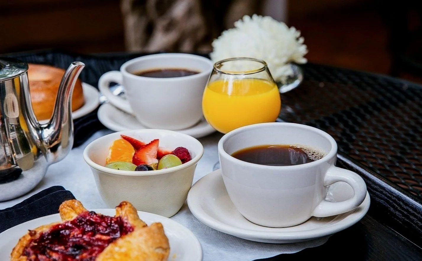 Breakfast table with cups of coffee, a glass of orange juice, a bowl of mixed fruit, a pastry, and a silver teapot.
