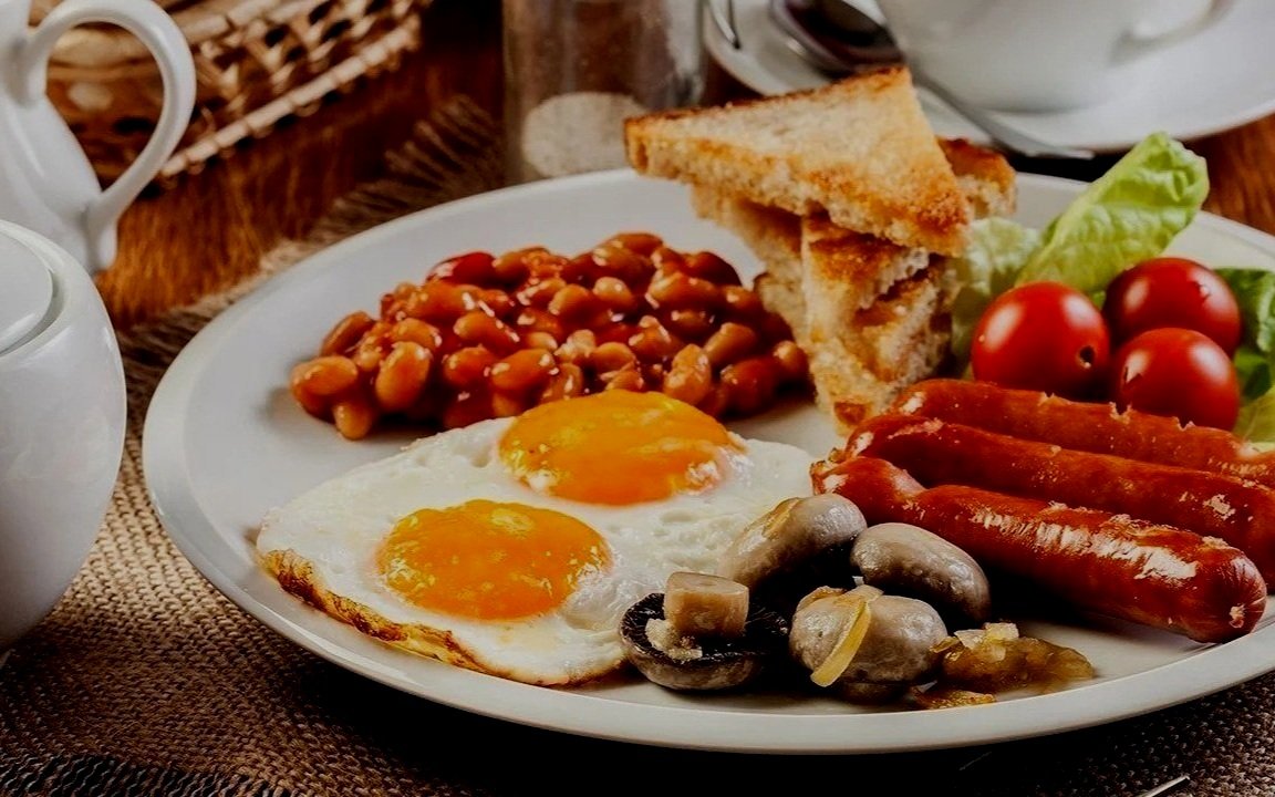 Plate with fried eggs, toast, sausages, baked beans, mushrooms, and cherry tomatoes.