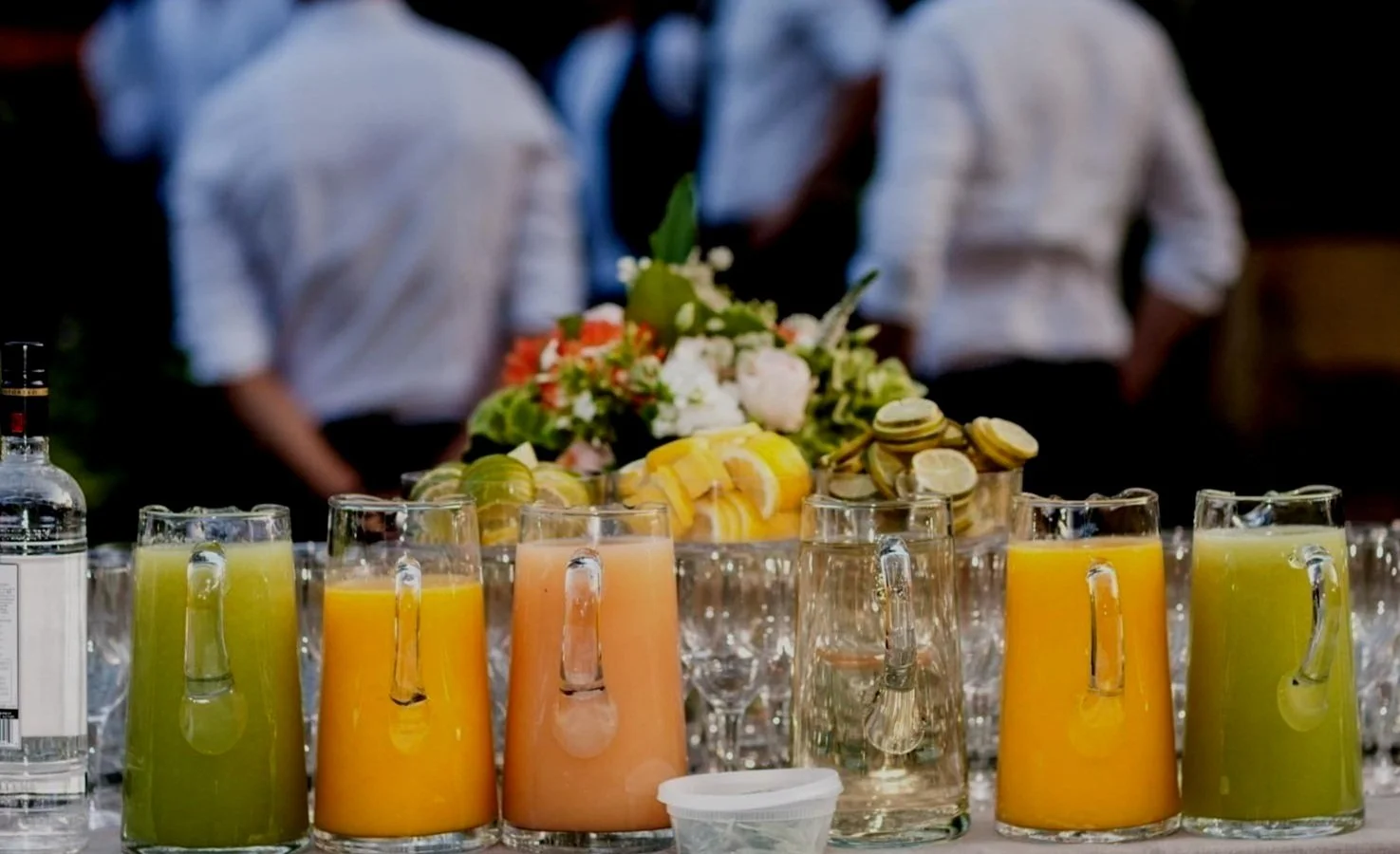 Table with pitchers of assorted juices, lemon and lime slices, floral arrangement, and bottles; people in background.