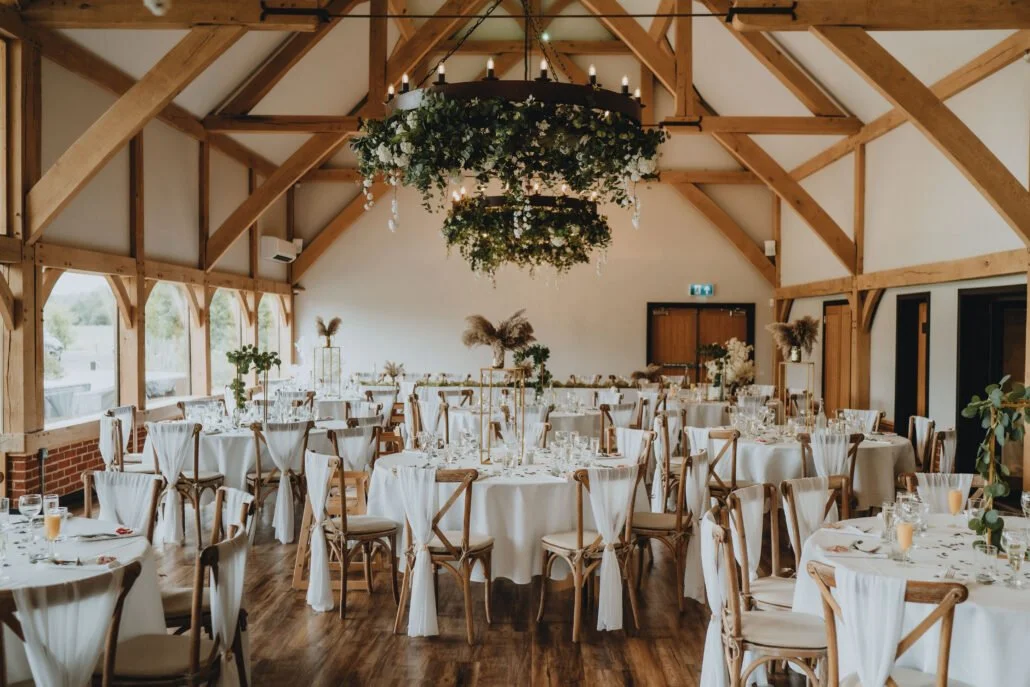 Wedding reception hall with round tables covered in white tablecloths, decorated with floral centerpieces and glassware, and surrounded by wooden chairs with white drapes. A large chandelier with candles and greenery hangs from the ceiling, and natural light streams in through the windows.