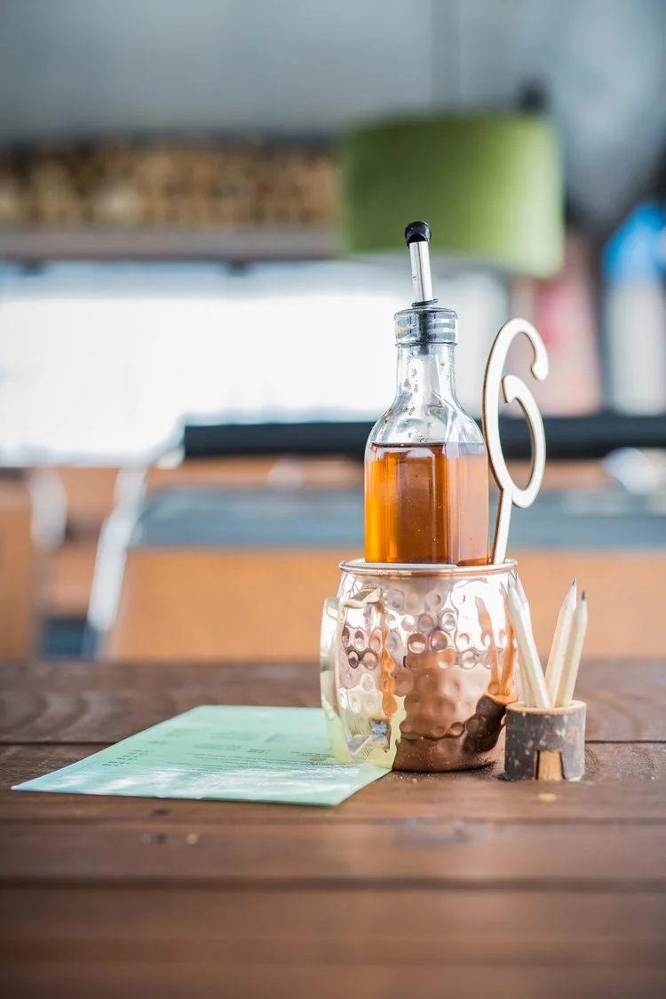 A wooden table setup with a glass bottle of oil or vinegar in a metal holder, alongside a small container with pencils.