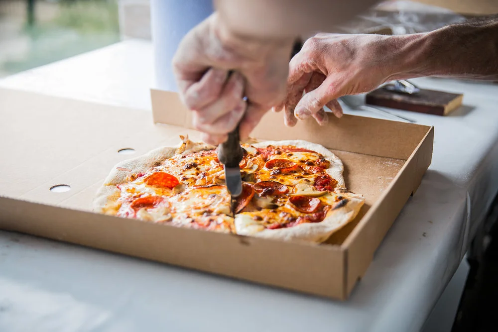 Person slicing a pepperoni pizza on a white table, with the pizza placed inside a cardboard box.
