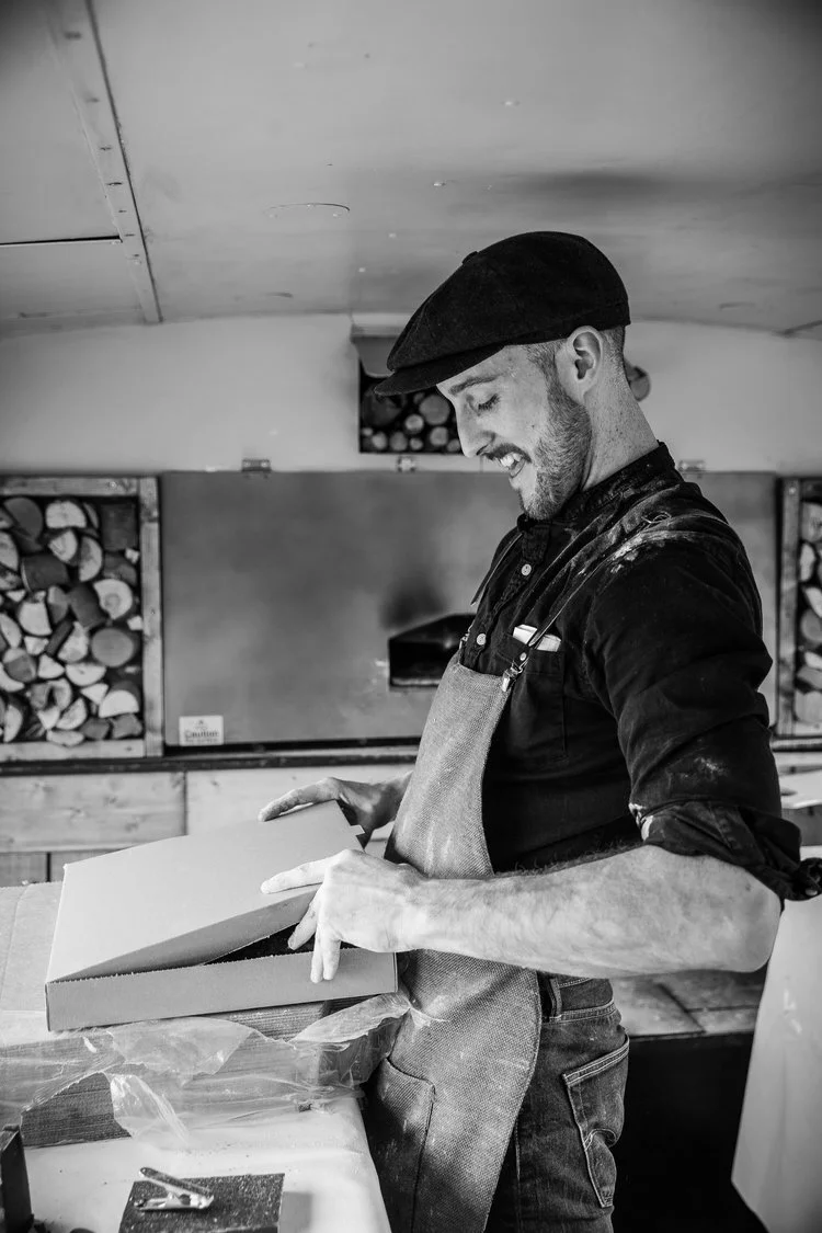 Man wearing an apron and hat, smiling, closing a pizza box, with wood-stacked oven in background.