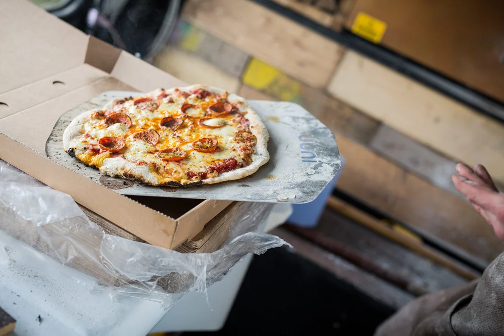 A freshly baked pepperoni pizza on a metal pizza peel next to an open pizza box.