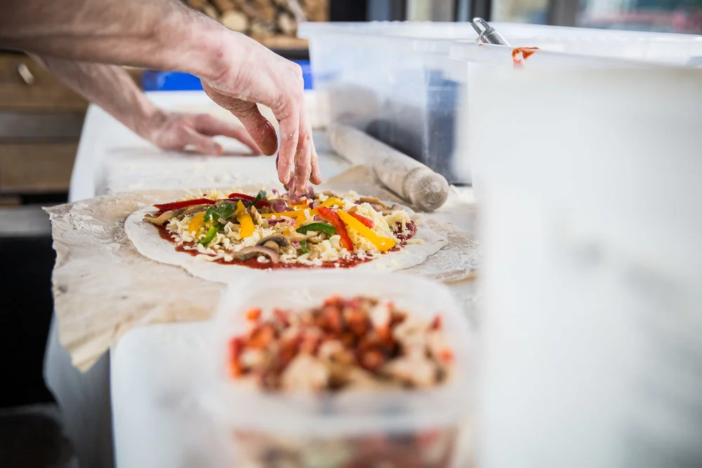 Person preparing a homemade pizza with vegetables and cheese on a marble surface.
