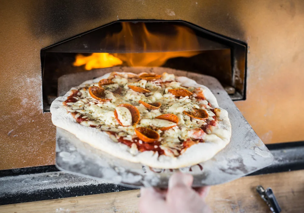 A handmade pizza with pepperoni and cheese being placed into a wood-fired oven.