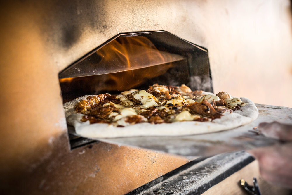 Pizza being placed into a wood-fired oven on a pizza peel with flames visible inside.