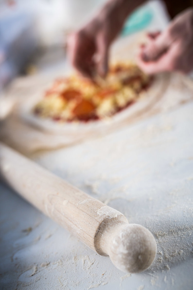 Close-up of a rolling pin on a floured surface with hands preparing a pizza in the background.